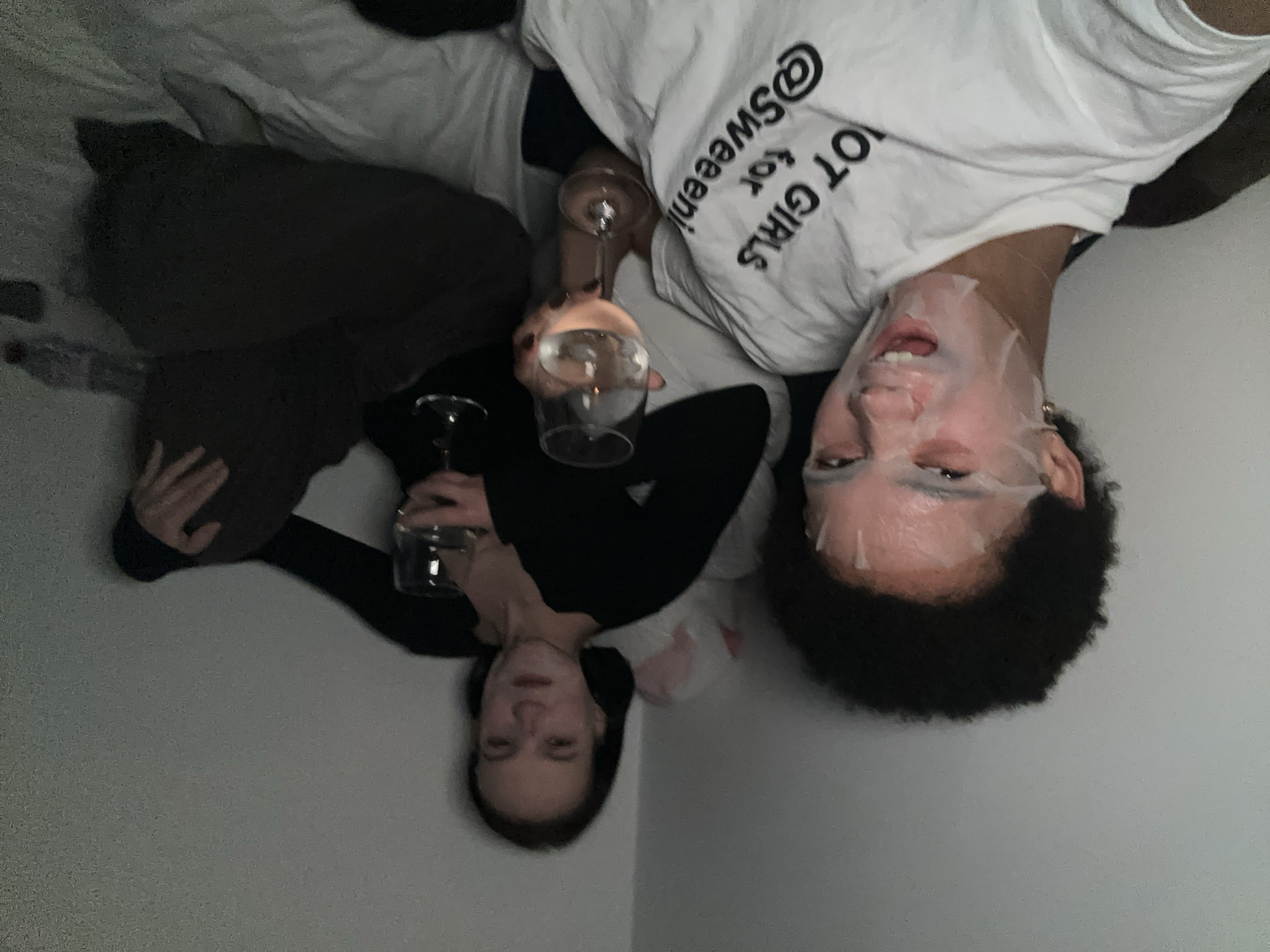The image shows two young women relaxing in a dimly lit room, likely a bedroom. The woman in the foreground has a sheet mask on her face and is wearing a white t-shirt with black text. She is smiling with her mouth open. The woman in the background is wearing a black top and dark pants, holding a glass of white wine. The room appears to be sparsely decorated, with a white wall and a bed. The overall mood is casual and relaxed, suggesting a night in. There is no indication of the specific location within London, United Kingdom.