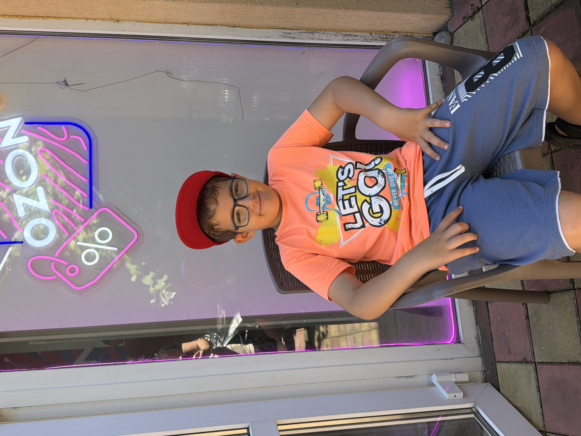 A young boy is depicted in a well-lit, upright medium shot, seated comfortably on a dark grey plastic chair next to a large glass window or door. He is wearing a red cap, black-framed glasses, a vibrant orange t-shirt, and blue shorts with white striped accents. The orange t-shirt prominently features the words "LET'S GO!" with a skateboard graphic and "NEVER GIVE UP" printed below. The boy looks directly at the camera with a relaxed expression, displaying a slight, pleasant smile, with his hands resting on his thighs.
Behind him, through the glass, illuminated neon signs are visible. One sign clearly spells "OZON" in white neon with a blue outline on part of the letter 'N'. Below it, a pink neon outline of a price tag with a white percentage symbol ("%") inside is displayed. Faint reflections of outdoor foliage and what appears to be another person's arm or hand can be seen in the lower section of the glass.
The setting suggests an outdoor commercial area or the entrance to a shop, typical of a street or plaza. The ground is paved with square tiles in various shades of grey, red, and brown. Above the window, a light brown building wall is visible. The bright lighting indicates it is daytime with clear weather. This scene captures a casual moment in a commercial area of Sochi, Russia.