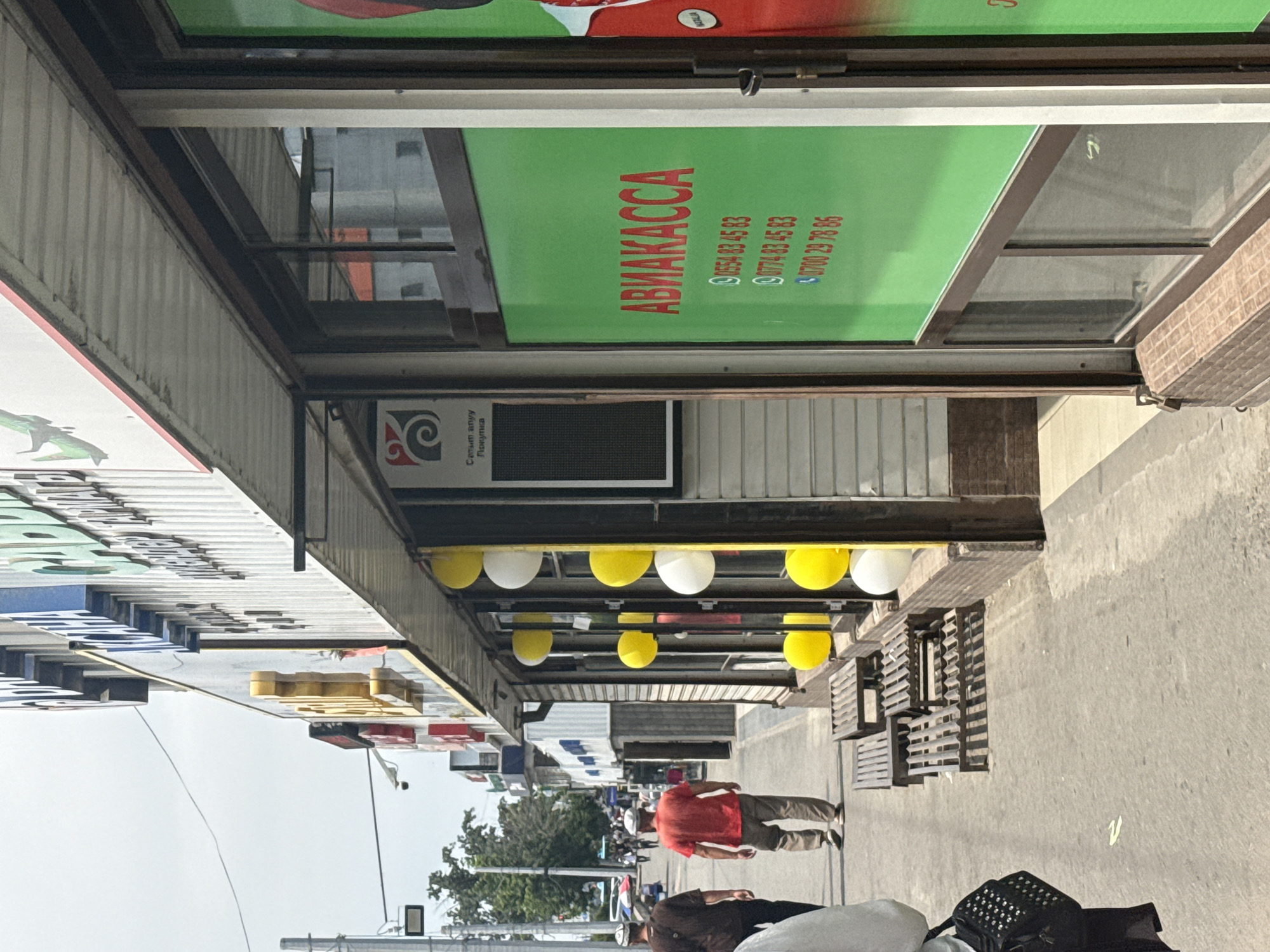 The image depicts an outdoor scene, likely a street or market, with buildings lining one side of the frame. The perspective is unusual, as the camera seems to be tilted or the image is rotated. In the foreground, a person wearing a red shirt and light brown pants is walking away from the camera. They appear to be casually strolling. Further down the street, other individuals are visible, suggesting a public space with some pedestrian activity.
Above the walkway, several shops or businesses are present. One prominent sign displays the word "АВИАКАССА" in large red letters against a vibrant green background. Below this text, there are several phone numbers listed: "0554 83 45 83", "0774 83 45 83", and "0700 29 78 86". Another sign to the left, partially visible, contains Cyrillic text and a stylized logo. Decorative yellow and white balloons are suspended above the storefronts, adding a festive or promotional touch.
The setting appears to be a commercial area with a pedestrian walkway and storefronts. The time of day seems to be daytime, judging by the natural light illuminating the scene. The weather appears to be fair, with no indication of rain or adverse conditions. The overall atmosphere is that of a busy, everyday environment. The image does not convey strong emotions, rather it documents a slice of urban life.