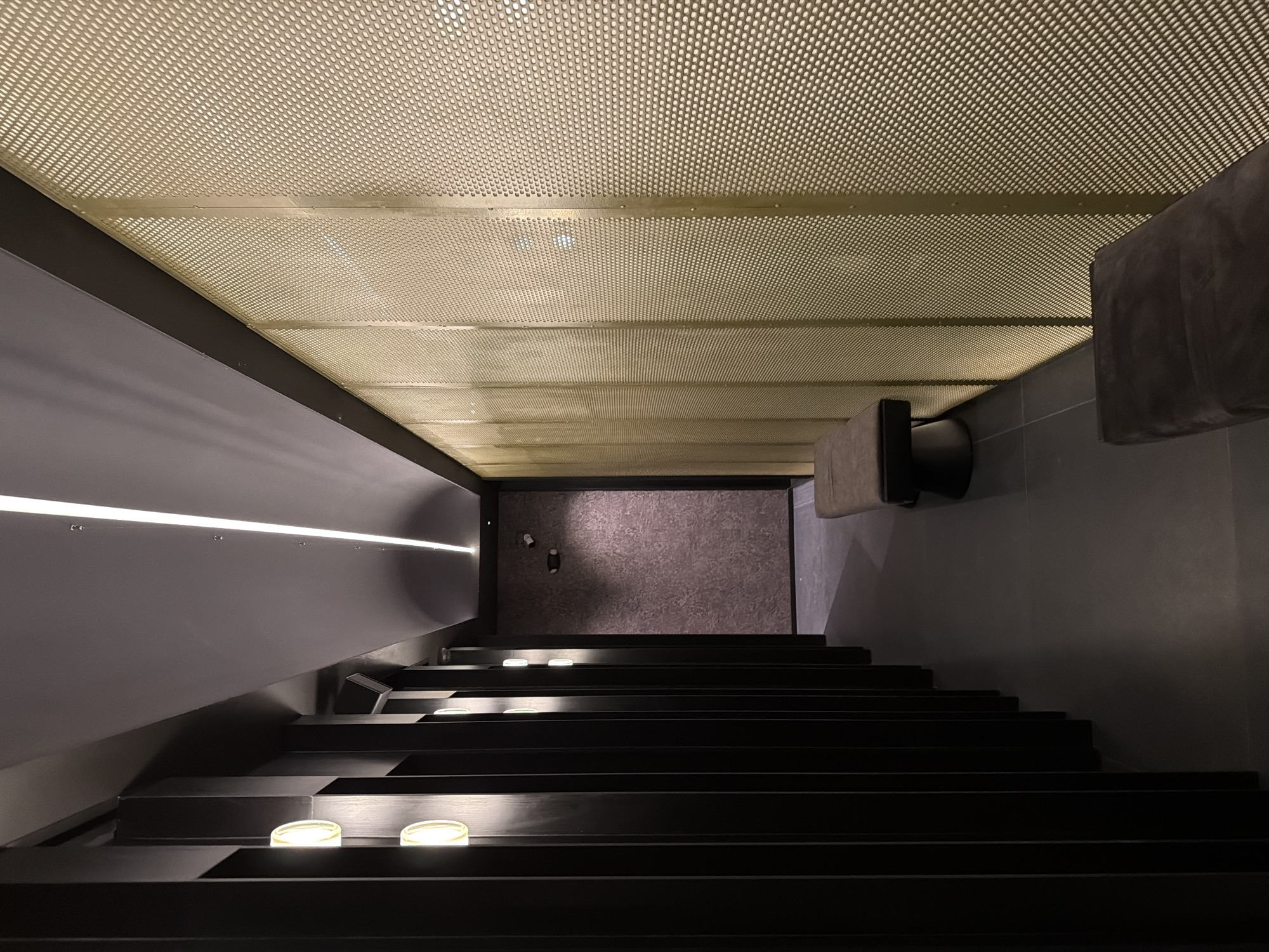 A view looking down a set of dark, modern stairs toward a wall at the bottom of the stairwell. The stairs have small lights recessed into the risers. A linear light fixture is visible on the wall to the left of the stairs. The ceiling above the stairs is a perforated metal grid. The wall to the right of the stairs is dark gray. The location is purportedly San Bernardino Tlaxcalancingo, Mexico.