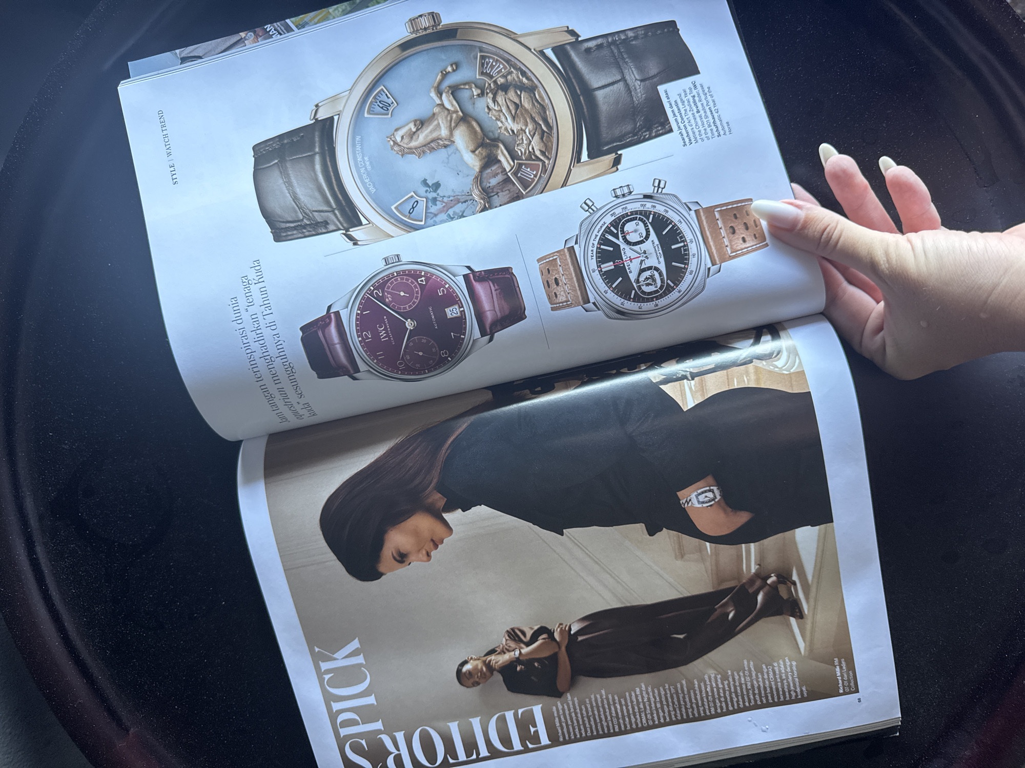 A close-up overhead shot captures a person's manicured hand holding open a magazine, displaying articles about luxury watches. The magazine lies on a dark, textured surface, possibly a car dashboard or a table, with bright natural light illuminating the pages. The setting appears to be Kuta Selatan, Indonesia, given the provided location context.
The top page of the magazine features three distinct high-end watches:
1. A Vacheron Constantin Métiers d'Art timepiece, prominently displayed, showcasing an intricate horse sculpture on its dial, accompanied by text mentioning "VACHERON CONSTANTIN GENEVE" and "YEAR OF THE HORSE."
2. An IWC Schaffhausen Portugieser Automatic watch with a deep purple dial and leather strap.
3. A Breitling Top Time B01 Shadow Rider chronograph with a black dial and a light brown perforated strap.
Accompanying text on this page includes "STYLE / WATCH TREND" and descriptions in what appears to be Indonesian, with phrases like "Jam tangan terinspirasi dunia equestrian menghadirkan 'tenaga kuda' sesungguhnya di Tahun Kuda" (Horse-inspired watches bring true 'horse power' to the Year of the Horse). English captions specify watch models like "Vacheron Constantin Métiers d'Art The Legend of the Chinese Zodiac Year of the Horse," "Breitling Top Time B01 Shadow Rider," and "IWC Schaffhausen Portugieser Automatic 42 Year of the Horse."
The bottom page features an "EDITOR'S PICK" section with two female models wearing Richard Mille watches. One model, dressed in a dark outfit, stands, while another, in a brown ensemble, sits or crouches. Visible text describes details of Richard Mille watches, referencing models like "RM 037" and "RM 07-01," and mentioning features like "snow-set," "intergalactic," and various diamond settings. The overall impression is a showcase of luxury timepieces in a glossy magazine.