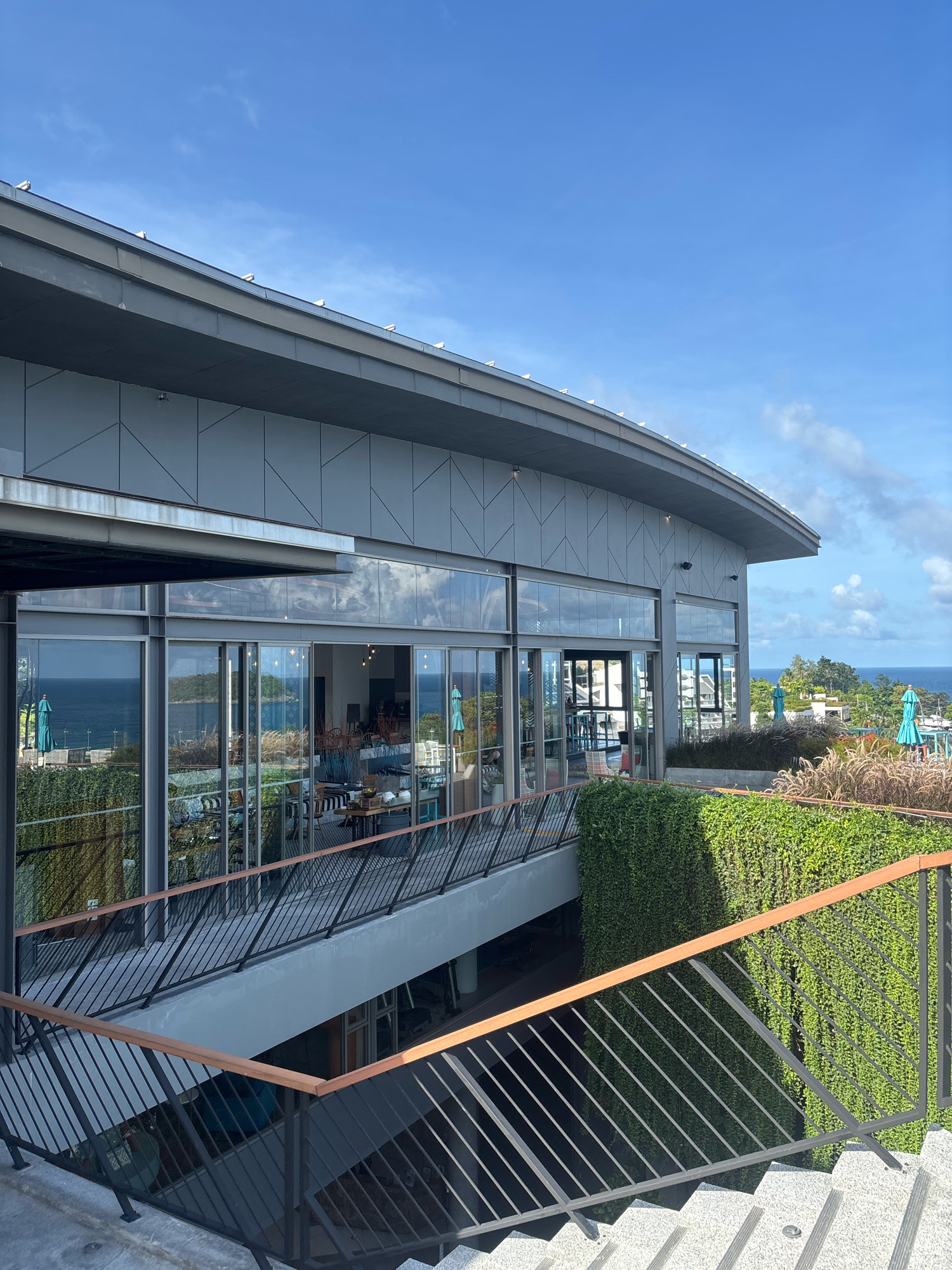 The image shows a multi-story building with a curved roof and large glass windows. The building overlooks the ocean. There are stairs and railings leading down to lower levels, and lush green vegetation covers parts of the building's exterior. Blue umbrellas are visible on an outdoor patio area. The sky is clear with a few wispy clouds. The scene appears to be a resort or restaurant in Karon, Thailand.