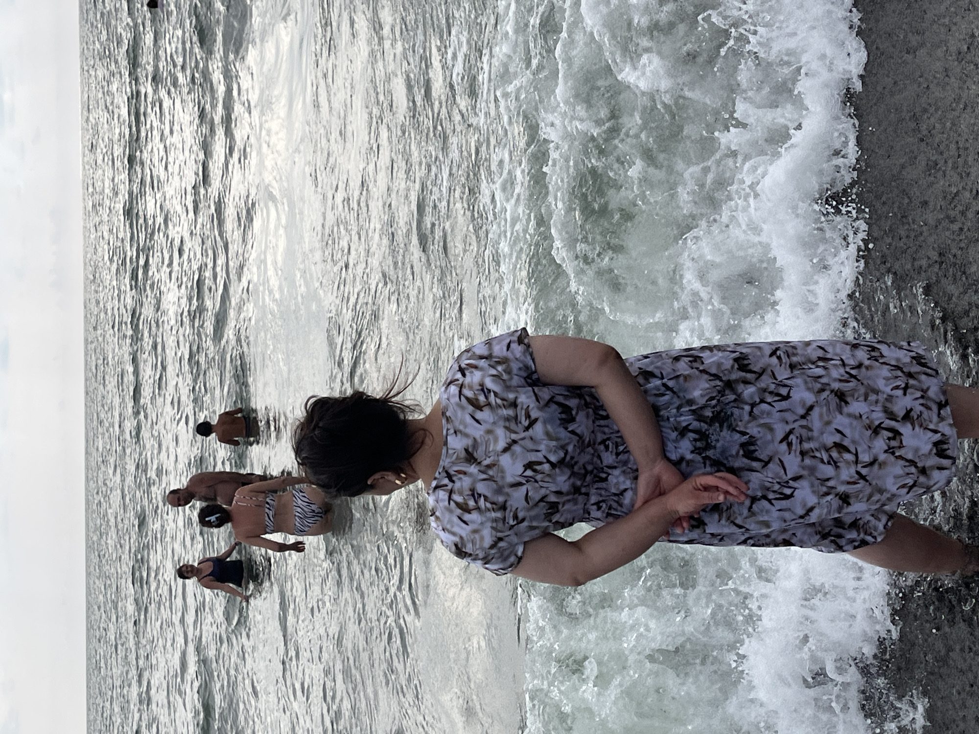The image shows a group of people in the sea, with a wave crashing on the shore in the foreground. A woman in a floral dress is standing in the water, facing away from the camera, with her hands clasped behind her back. Behind her, several other people are also in the water, some swimming and others standing. The sea is choppy, with white foam from the waves. The sky is overcast, suggesting it might be a cloudy day. The scene appears to be a beach or a coastal area, possibly during daytime given the light. The overall mood is relaxed and casual, with people enjoying the water. There is no visible text in the image. The location is Ureki, Georgia.