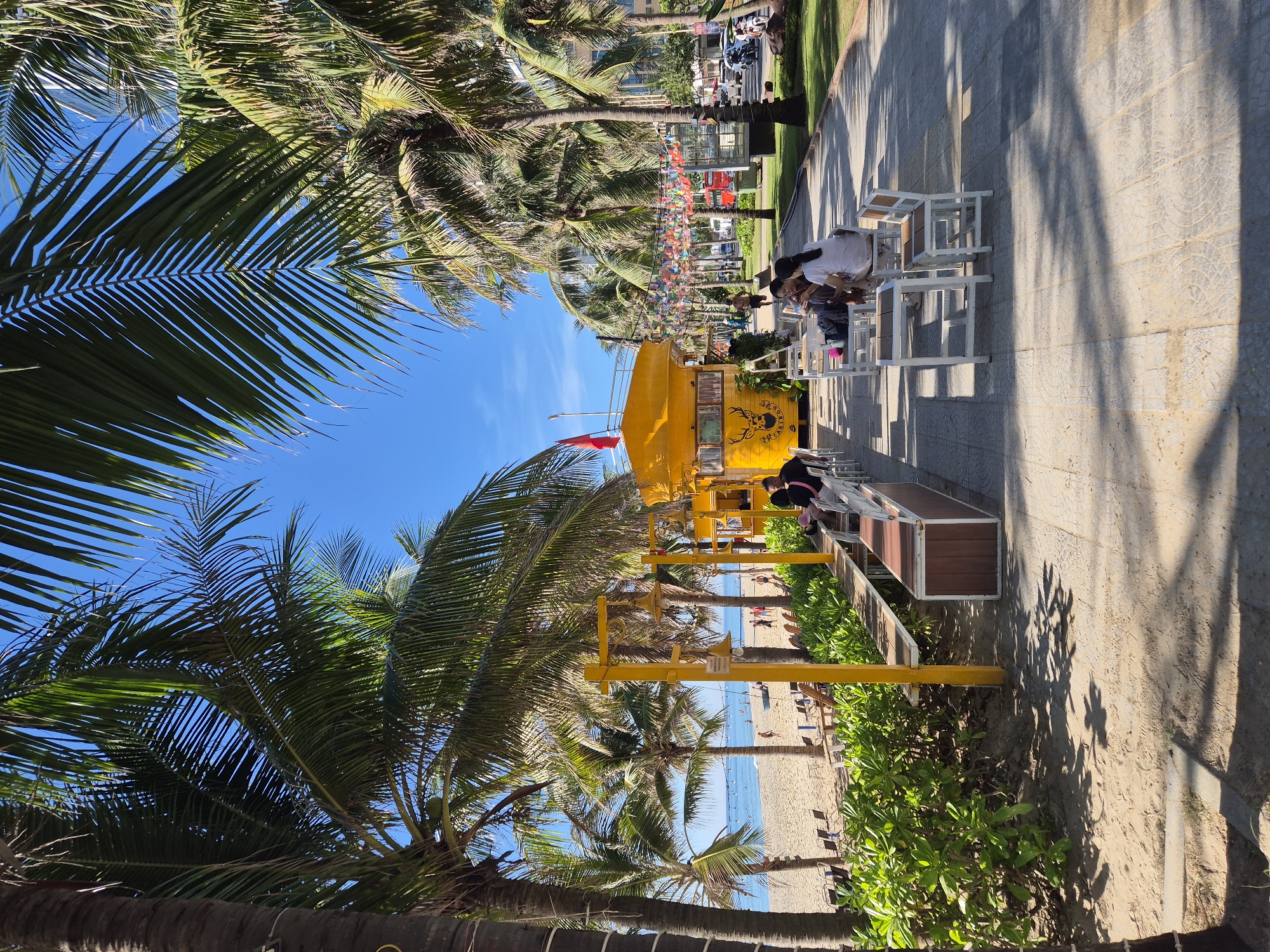 This is a full-shot, eye-level, outdoor photograph of a beachfront area in Da Nang, Vietnam. The scene is dominated by lush palm trees with green fronds and brown trunks, framing a clear blue sky. In the foreground, a sandy beach stretches towards the ocean, dotted with beach chairs and a few people. Adjacent to the beach is a paved walkway lined with more palm trees.
On the walkway, a vibrant yellow food truck with a rustic design, featuring a stylized deer logo and "THIGARINDO" text, is parked. Several tables and white chairs are set up in front of the food truck, where a few individuals are seated, some engaged in conversation. The walkway continues past the food truck, leading towards buildings and a street with vehicles in the background. A string of colorful international flags is suspended above the walkway between lampposts. The overall atmosphere is relaxed and tropical.