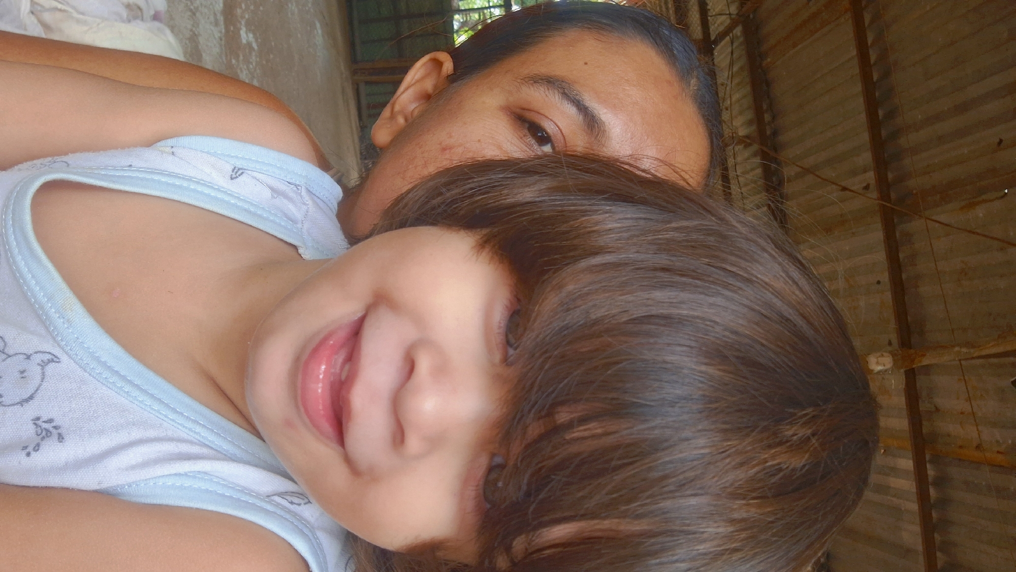 Two individuals are captured in a close-up, low-angle selfie. The foreground is dominated by the face of a young child, whose brown hair obscures much of their forehead and eyes, but their smiling mouth and rosy cheeks are clearly visible. The child is wearing a light blue tank top with a white trim and a subtle pattern. Behind the child, partially obscured by their head and hair, is an adult whose face is turned to the side, with their right eye visible and a portion of their ear. The adult appears to be looking towards the camera. The background consists of a rustic, corrugated metal roof, suggesting an outdoor or semi-outdoor setting. The overall lighting is warm and natural. The image appears to have been taken in Vallecito, Venezuela, due to the style of the roof and the ambient conditions which are consistent with the region.