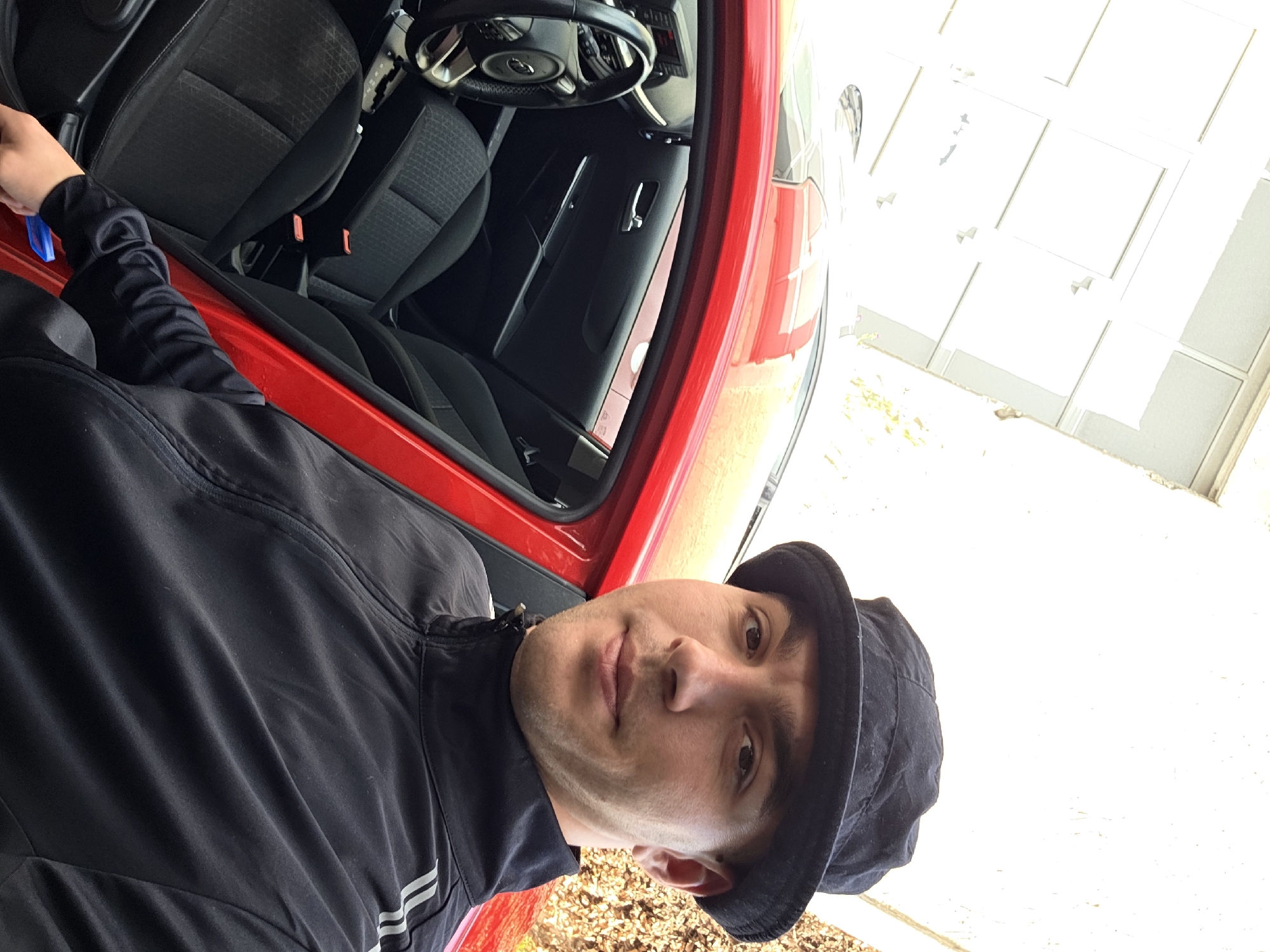 A young man wearing a black bucket hat and a black zip-up jacket stands next to the open door of a red car. The car's interior, with black seats and a steering wheel, is visible. The man is looking directly at the camera and is holding a blue object in his right hand. The scene appears to be outdoors, with a white wall and some foliage in the background.