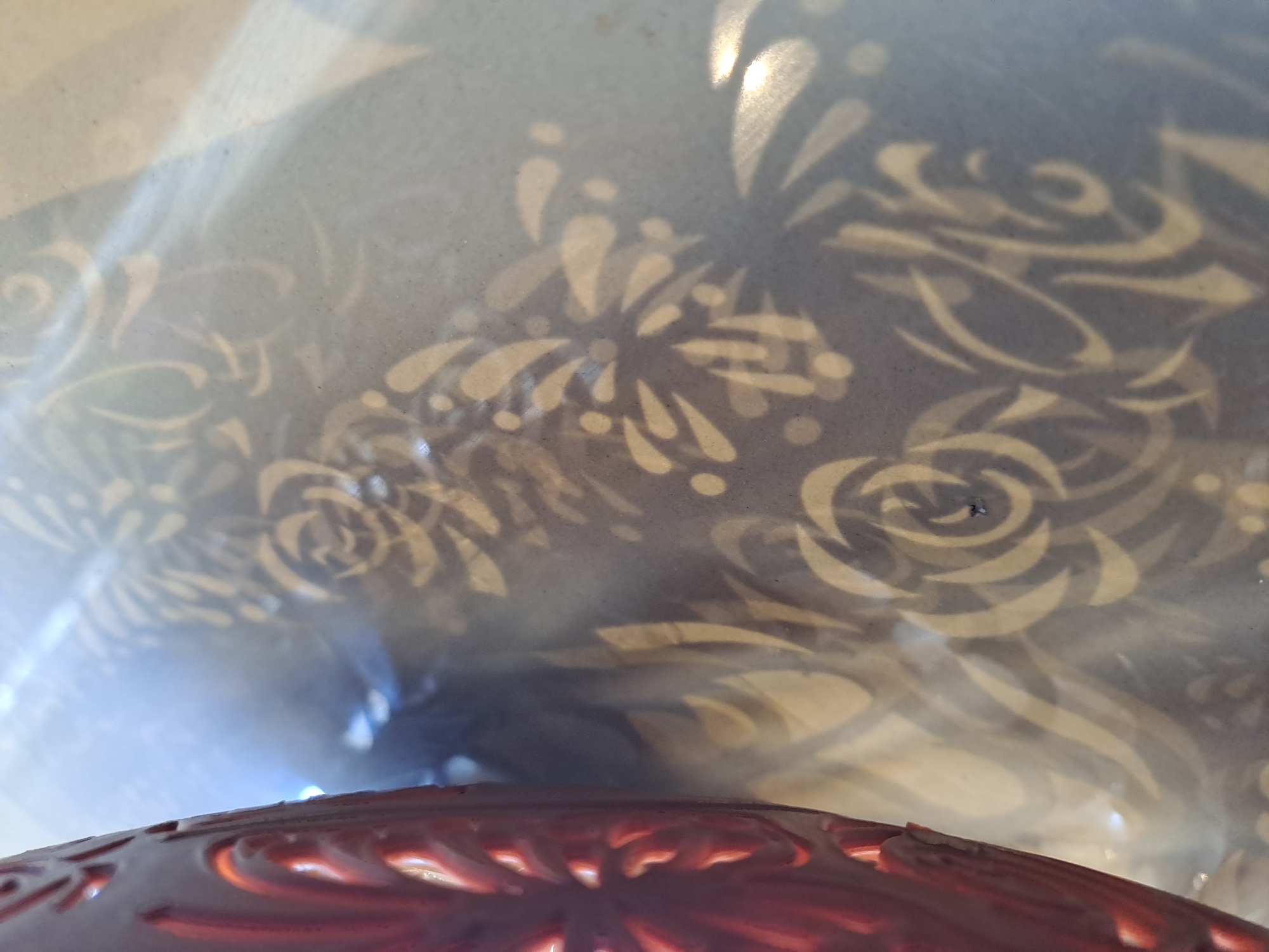 The image is a close-up, overhead shot of a patterned object casting shadows on a surface. The object itself, visible at the top, appears to be a deep red or maroon color with intricate cut-out or embossed designs that resemble floral or swirling motifs. The shadows cast by this object are a lighter beige or cream color and form repeating patterns of stylized flowers and curves. These shadows are projected onto a gray surface, possibly a table or a floor, which has a slightly mottled texture and some shiny, reflective areas. The overall impression is one of light and shadow play, highlighting the decorative elements of the object. The image does not depict any specific location in Bireuenmeunasahdayah, Indonesia, nor does it contain any people, actions, or interactions.