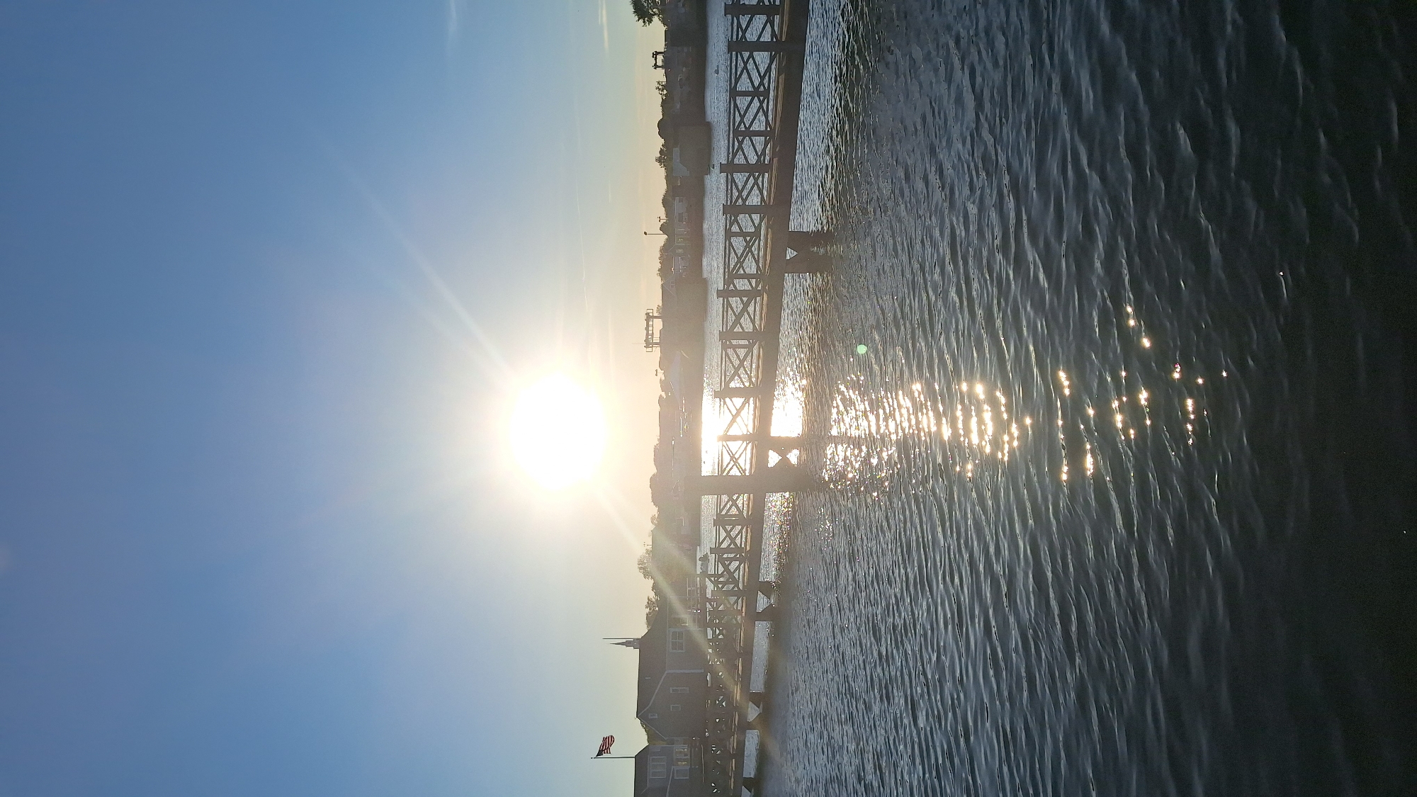 This image depicts a serene waterside scene in Nantucket, Massachusetts, United States of America. The sun, prominently positioned in the sky, casts a bright glare and creates a shimmering reflection on the rippling water. A wooden pier extends across the frame, featuring a decorative railing with a crisscross pattern. On the left side of the pier, a building with a red-roofed section and a white spire is visible, alongside an American flag flying from a flagpole. The background shows a line of structures and trees, suggesting a coastal town or harbor area. The water appears calm, with gentle waves catching the sunlight. The overall atmosphere is tranquil and bathed in the warm light of the sun.