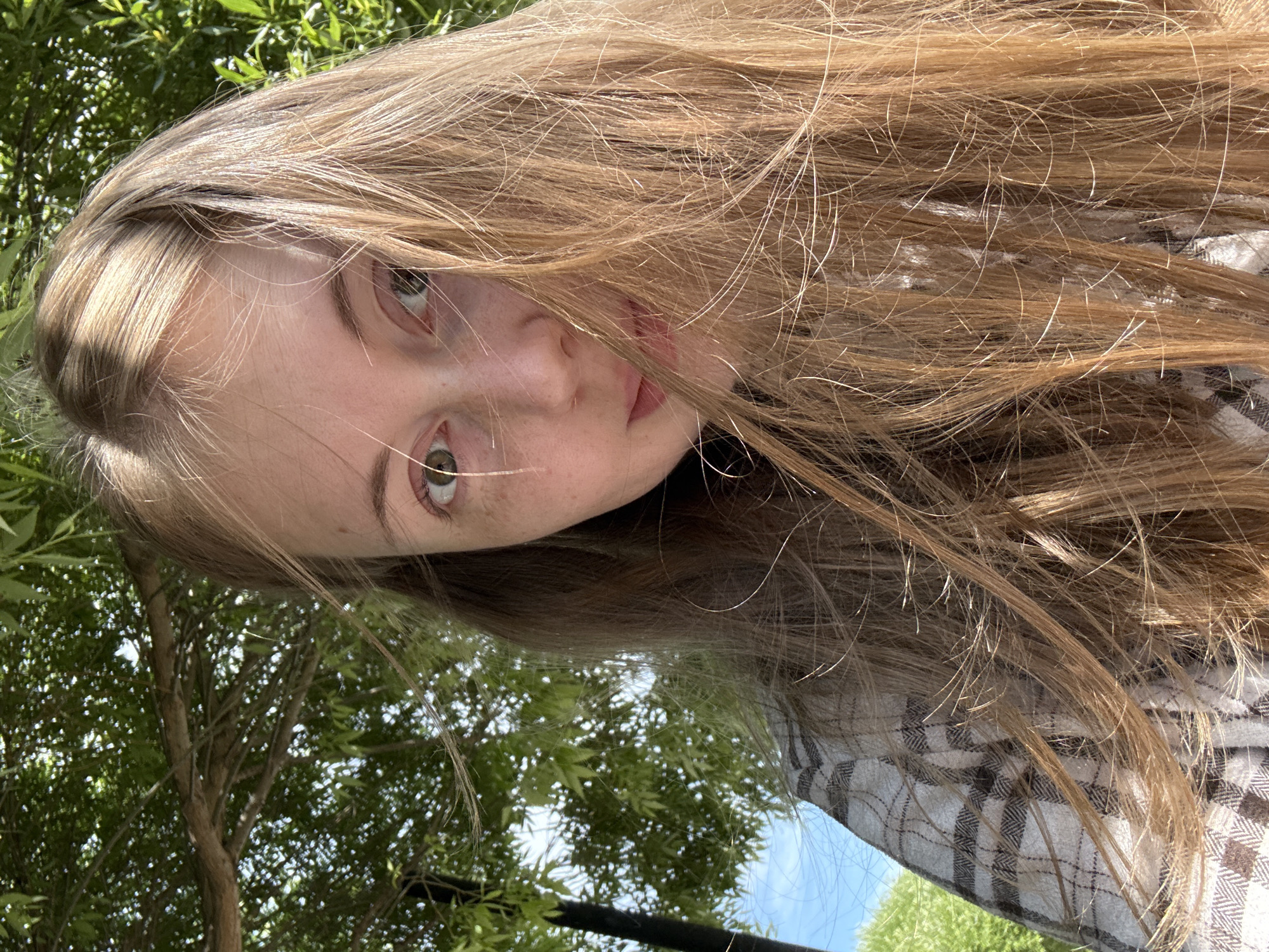 This is a close-up, slightly tilted photograph of a young person's face, with their long, light brown hair falling across the frame. The subject's eyes are open and looking towards the camera, with a soft, natural expression. Sunlight catches the strands of their hair, highlighting lighter tones and creating a slightly windswept appearance. A portion of their face, including an eye, cheek, and lips, is visible. They are wearing a plaid shirt.
The background is blurred but suggests an outdoor setting with lush green foliage and a bright blue sky. The lighting indicates it is daytime, likely sunny. The overall impression is casual and relaxed. There is no visible text in the image.