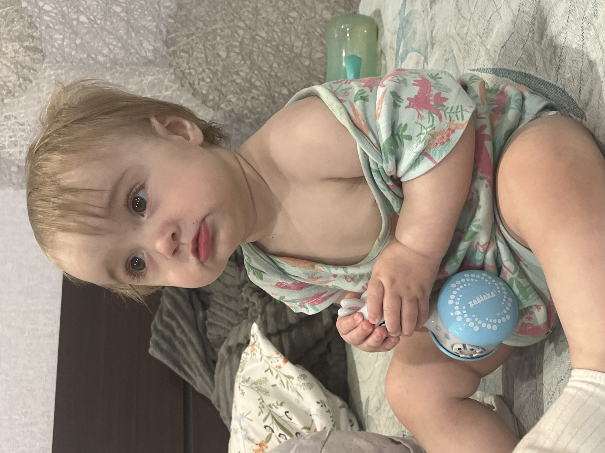 A child sits on a bed, holding a blue toy with white dots. The child wears a light blue outfit with pink and orange designs. A green bottle is visible behind the child. The image was taken in a location of Chaikovsky, Russia.