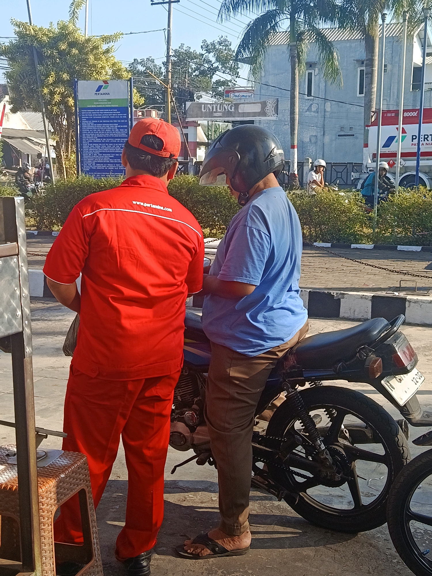 Two individuals stand next to a parked motorcycle on a paved area, likely a street or parking lot, in Tempeh Lor, Indonesia. The person on the left wears a bright red uniform with a red cap, standing facing away from the camera. A white logo with the text "www.pertamina.com" is visible on the back of their shirt. The person on the right is seated on the motorcycle, wearing a blue t-shirt and brown pants, with a black helmet. The motorcycle is dark colored, with a visible license plate, and appears to be parked adjacent to a low hedge. In the background, a Pertamina gas station sign is prominent, featuring the Pertamina logo and text in Indonesian. Various other buildings, palm trees, and street infrastructure, including a fuel truck, are visible in the distance, suggesting an urban or semi-urban environment. The lighting indicates it is daytime, with sunlight casting shadows.