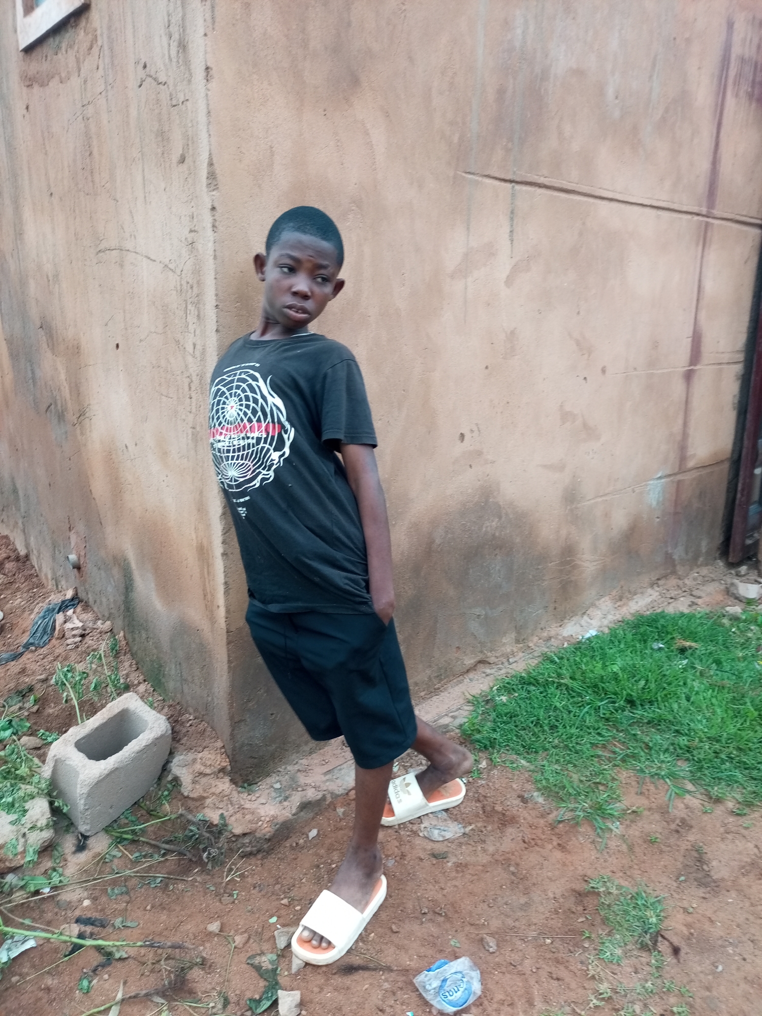 A young Black boy stands near a textured, light brown wall in Tempure, Nigeria. He is wearing a black t-shirt with a white circular graphic and black shorts. On his feet are white flip-flops. He is leaning against the wall with his right hand in his pocket, and his left arm is bent upwards. The ground is a mixture of dirt and patches of green grass. To the left of the boy, a hollow concrete block lies on the ground amongst some sparse vegetation. In the background, a dark, weathered metal doorframe is visible. The lighting suggests an overcast day.