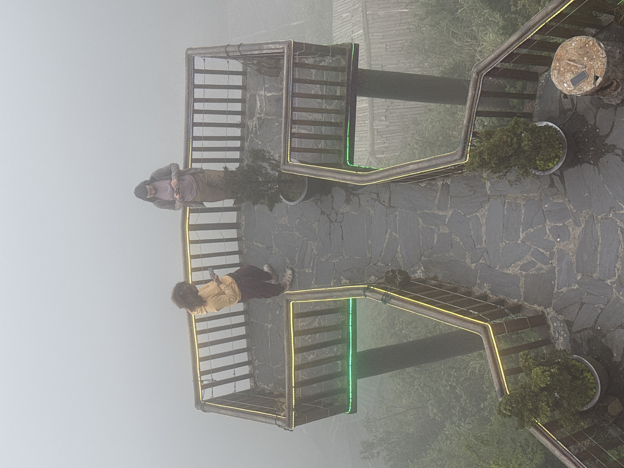 The image shows an elevated platform in Binh Lu, Vietnam, viewed from above and rotated counter-clockwise. The platform consists of two square structures with railings, interconnected by a walkway. The railings are illuminated with yellow lights, and one side of one structure is illuminated with green lights. Two individuals are present on the platform; one is wearing a tan jacket and dark pants, and the other is wearing a gray jacket and dark pants. Both appear to be looking at or using their phones. The ground surrounding the platform is paved with gray stone. Potted plants are situated at the corners of the platform. Beyond the platform is a dense fog, obscuring the surrounding environment. A tree stump or table is visible near one corner of the platform, on which a phone rests.