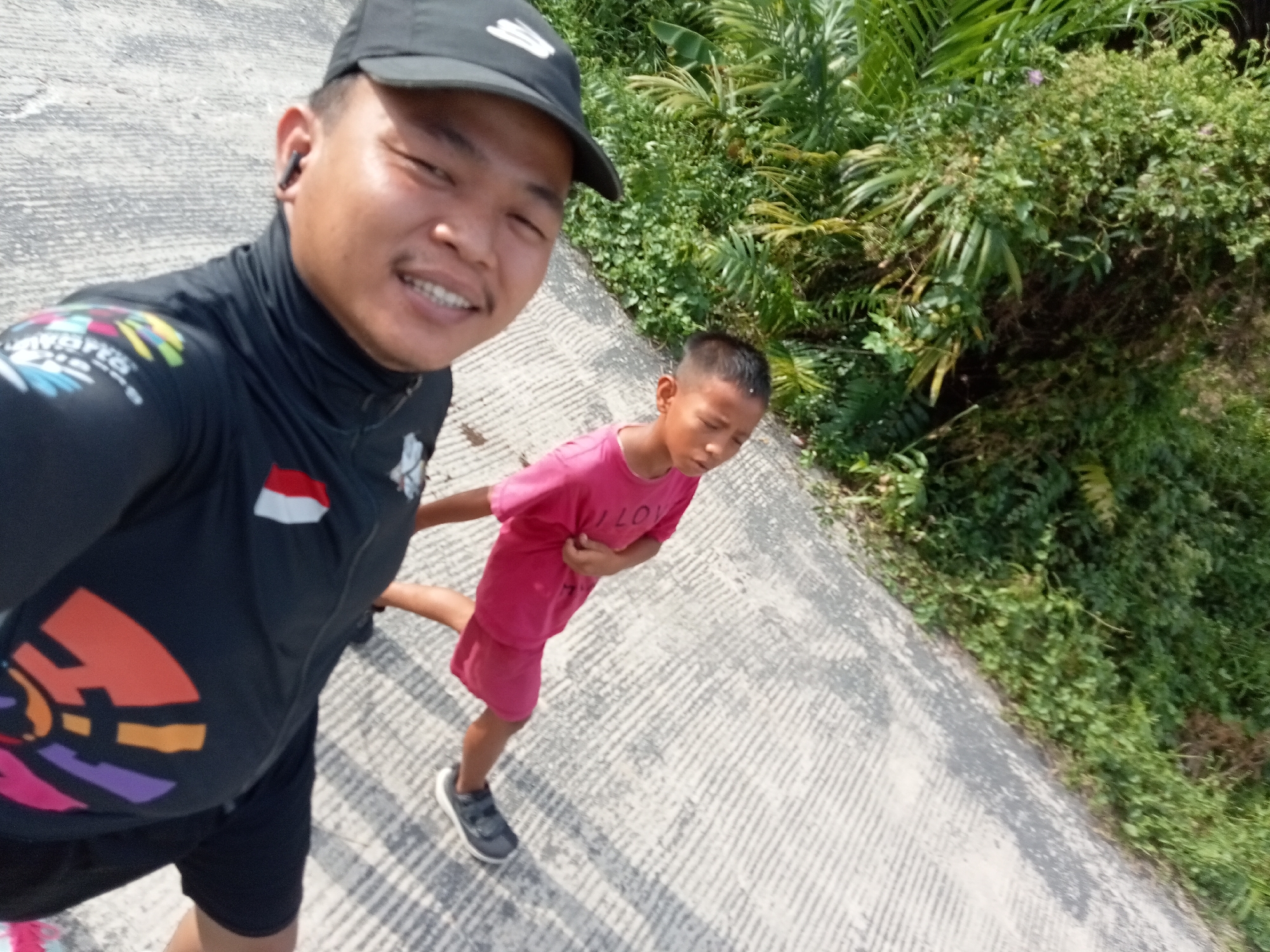 A man wearing a black baseball cap and a black athletic jacket is taking a selfie with a boy wearing a pink t-shirt and shorts. The man is smiling and has a mustache. The boy is looking down and has his left arm raised. They are standing on a concrete surface next to a lush green vegetated area. The concrete surface appears to be a path or walkway. The image is taken from a high angle, looking down at the subjects. The man is wearing wireless earbuds. The jacket has colorful graphics on the left chest and a small Indonesian flag on the right sleeve. The boy's t-shirt has text on it, but it is not clearly legible. The background is filled with dense green foliage, suggesting an outdoor environment possibly in Teluk Kenidai, Indonesia. The lighting suggests it is daytime.