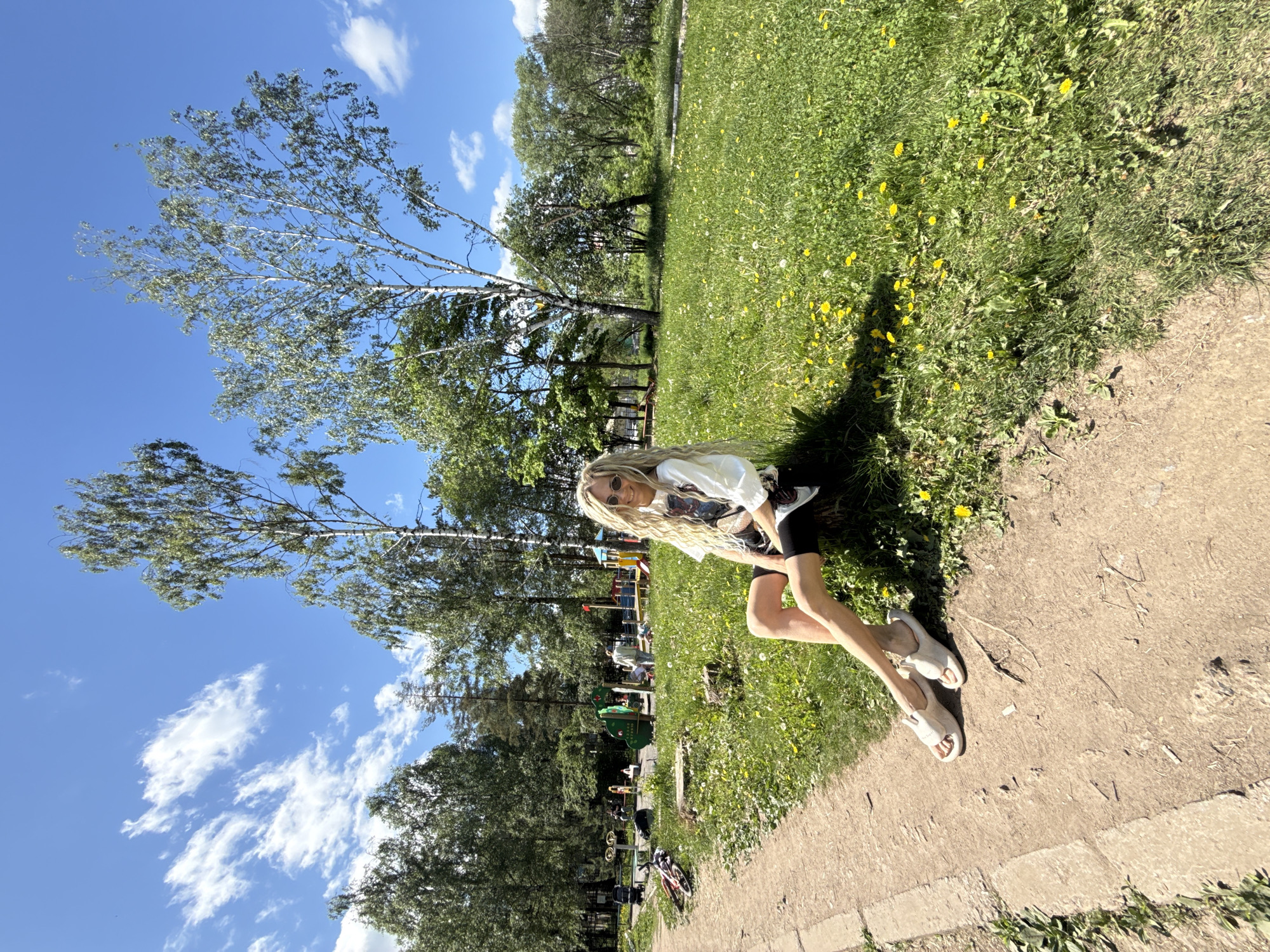 A woman is sitting on the edge of a concrete path in a park setting, likely in Rusanovo, Russia. The woman has long, blonde hair and is wearing sunglasses, a white T-shirt, and black shorts. She is wearing light-colored shoes. She is positioned on the right side of the image, with her legs extending onto the path. Behind her is a grassy area with trees, dandelions, and a playground. The sky is blue with scattered clouds. A bicycle is visible in the background.