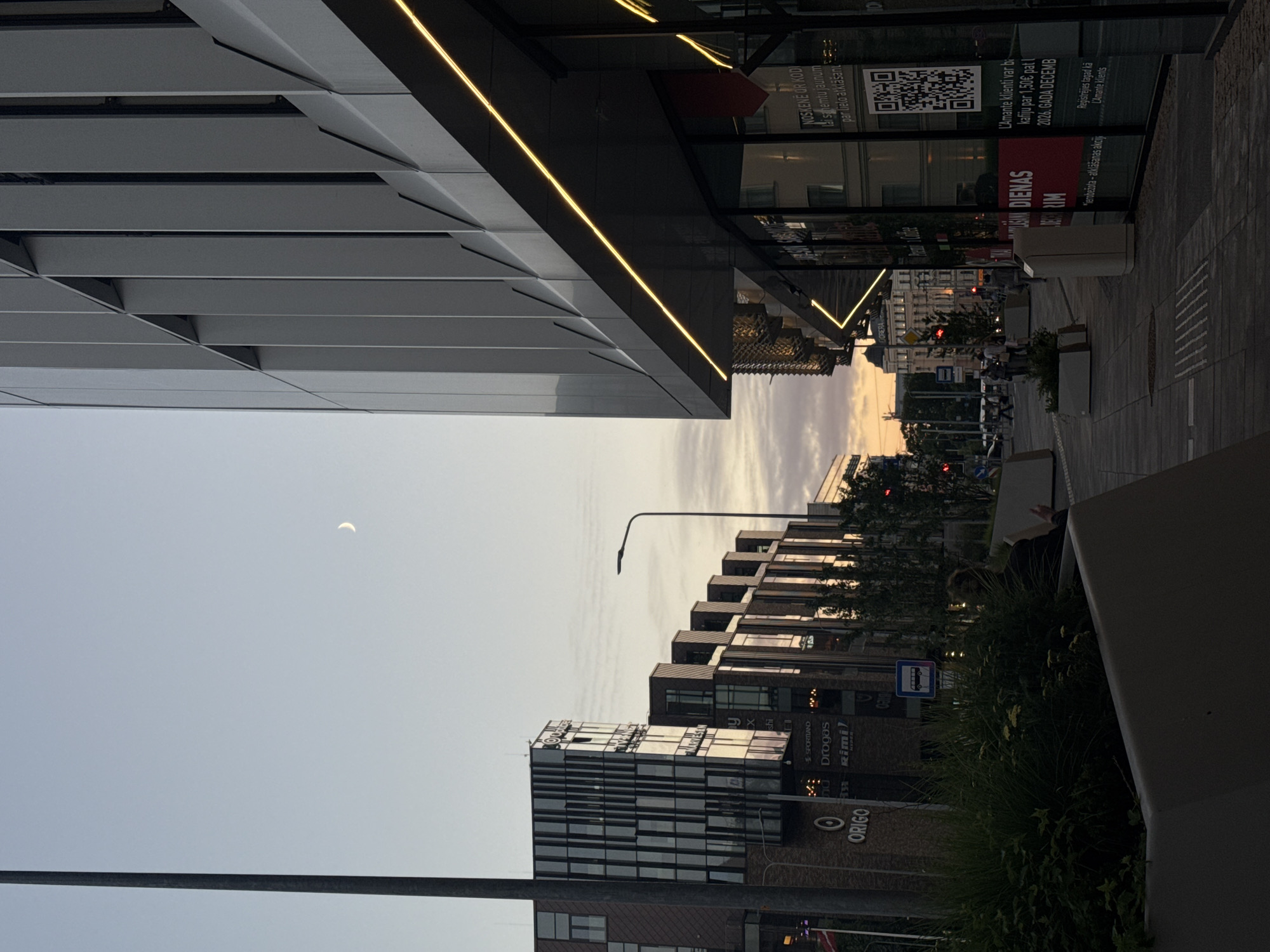 The image depicts a street scene in Riga, Latvia, at dusk. A modern, multi-story building with a facade of vertical gray panels dominates the right side of the frame, featuring prominent yellow LED lighting along its overhang. To the left, a row of buildings showcases a mix of architectural styles, including glass facades and dark brickwork, with the "ORIGO" logo visible on one of them. A tall, dark lamppost stands on the far left, partially obscuring the view of the buildings. The sky is a pale blue with wisps of clouds, and a crescent moon is visible. The street level features planters with greenery and a sidewalk made of paved stones. Several individuals are present on the sidewalk, appearing to walk or stand, though their features are not clearly discernible. Traffic lights are visible in the background, suggesting an active intersection. The overall impression is that of a commercial or mixed-use urban area in the early evening.