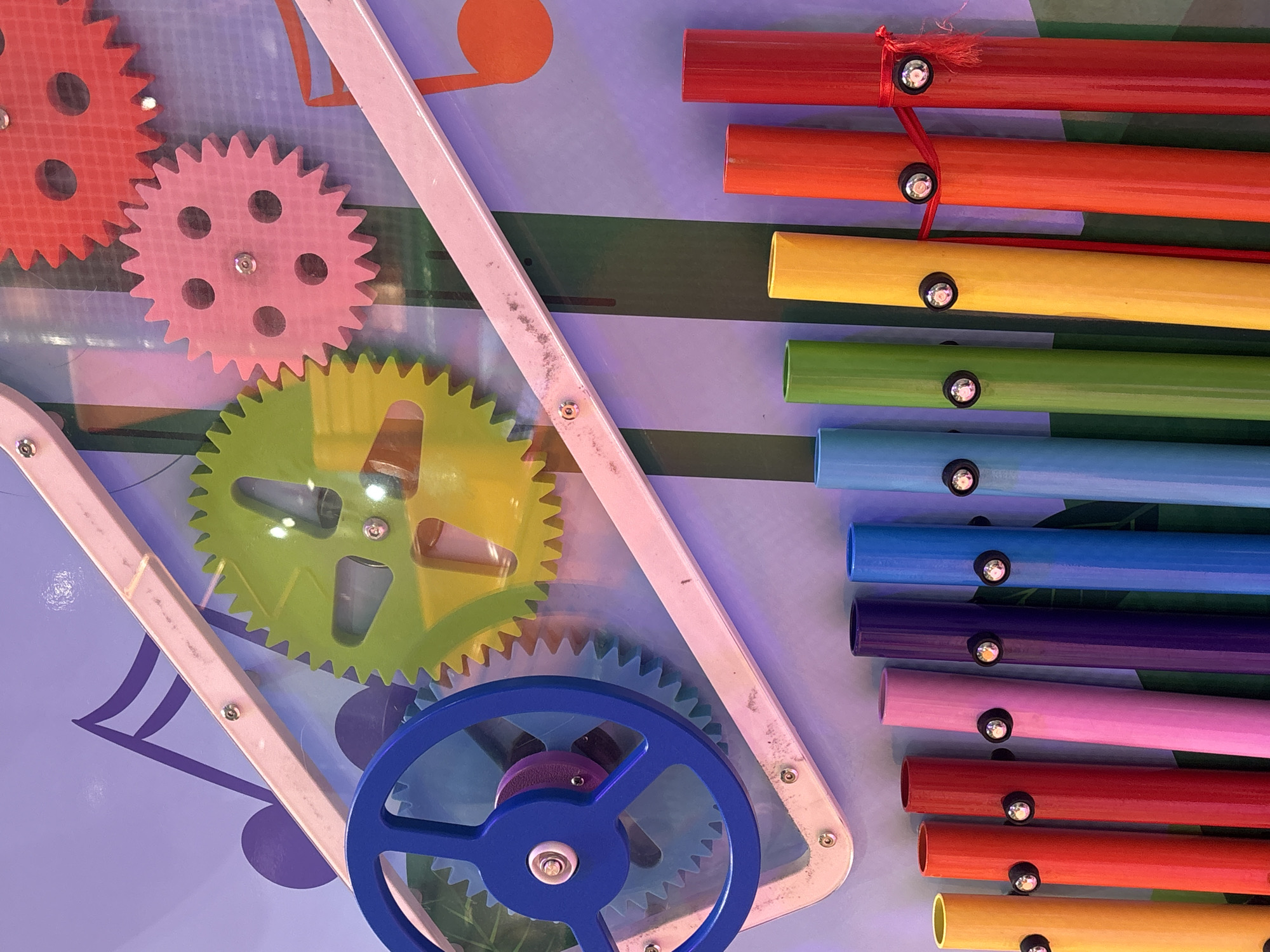 The image shows a colorful, child-friendly musical instrument, likely a xylophone with a gear mechanism. The instrument is mounted on a wall decorated with musical notes and vertical stripes in shades of purple and green. The xylophone features eight brightly colored tubes: yellow, red, red, pink, purple, blue, green, yellow, orange, and red, each with a black mallet holder secured by a screw. Some of the tubes are connected by a red string. Above the xylophone, a blue, green, pink, and red gear system is visible, suggesting interactive play. The lighting appears to be artificial and ambient, casting a soft glow on the scene. The image does not provide any specific visual cues to determine its location within the city of Dubai, United Arab Emirates.