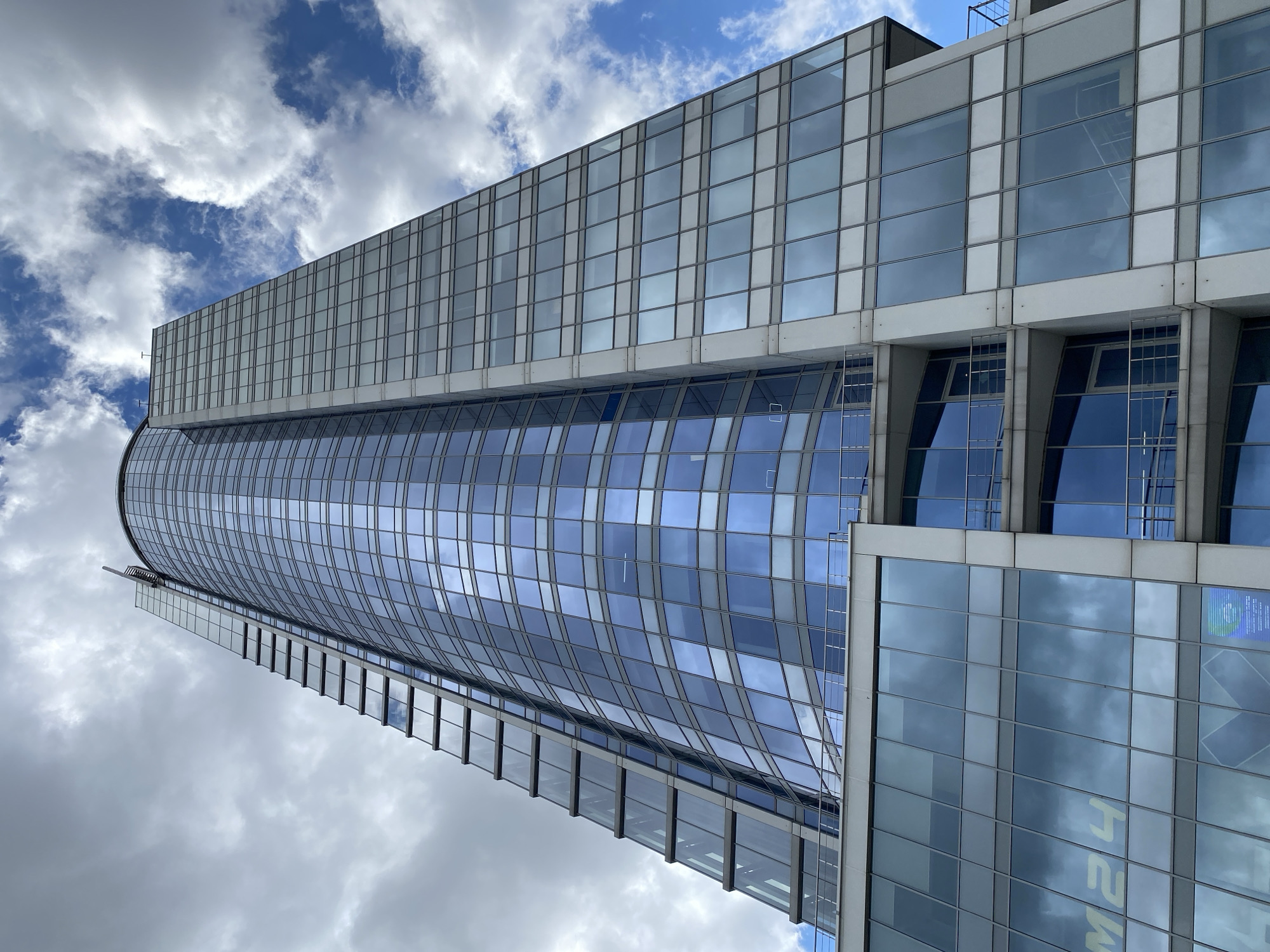 This is a low-angle, upward shot of a modern, curved glass skyscraper against a backdrop of a partly cloudy blue sky. The building features a sleek design with alternating bands of reflective blue glass and lighter-colored panels. The sky is dynamic, with fluffy white clouds scattered across the bright blue.
The building's facade is composed of numerous rectangular glass panes, reflecting the sky and surrounding clouds. A portion of the building on the right is more angular, with larger square glass panels. There appears to be a vertical element or antenna structure at the very top of the building.
The lighting suggests it is daytime, likely midday, given the brightness of the sky. The weather appears fair with some cloud cover. The scene evokes a sense of modern urban architecture and scale.
Visible text includes "M24" in large yellow letters reflected on the glass of the angular section of the building. There is also a small, partially visible blue and white sign on the same section, likely an advertisement or informational placard. The image's orientation is rotated 90 degrees counter-clockwise.