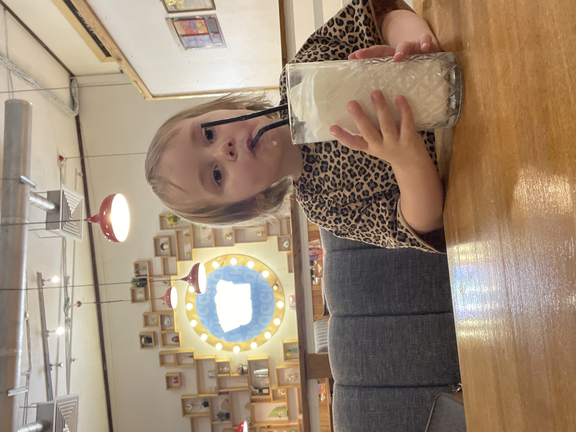 A young girl with blonde hair is seated at a wooden table in what appears to be a cafe or restaurant. She is wearing a leopard-print top and is holding a tall glass filled with a white, frothy drink, using two straws to sip it. The background shows a wall with shelves displaying various items and a large circular mirror with lights around its perimeter. Red pendant lights hang from the ceiling, and industrial-looking air ducts are visible. The overall setting suggests an indoor establishment, possibly a themed cafe, but the specific location within Vladivostok, Russia, cannot be determined from the visual information provided.