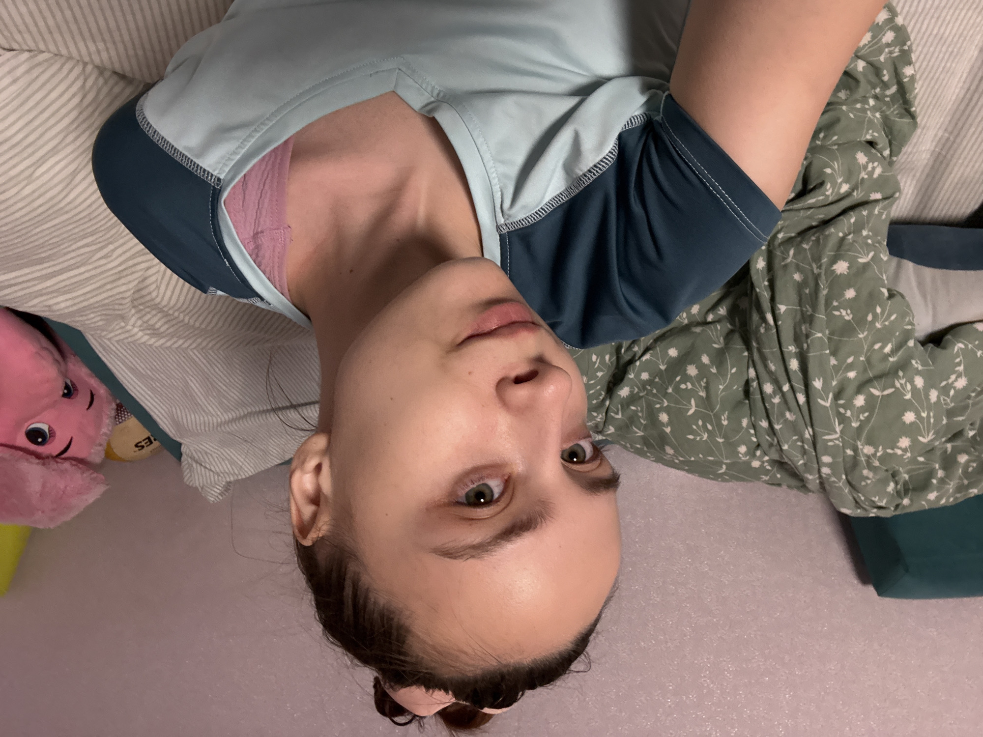 A woman lies on a bed covered with various bedding. She is wearing a light blue short-sleeved shirt with dark blue sleeves and a pink tank top underneath. The bedding includes a striped pillow, a green floral patterned duvet, and a teal pillow. A pink plush toy is visible on the left side of the image. The woman is looking directly at the camera. The background is a light purple sheet. The location, Прикамский, Russia is not discernible from the image.