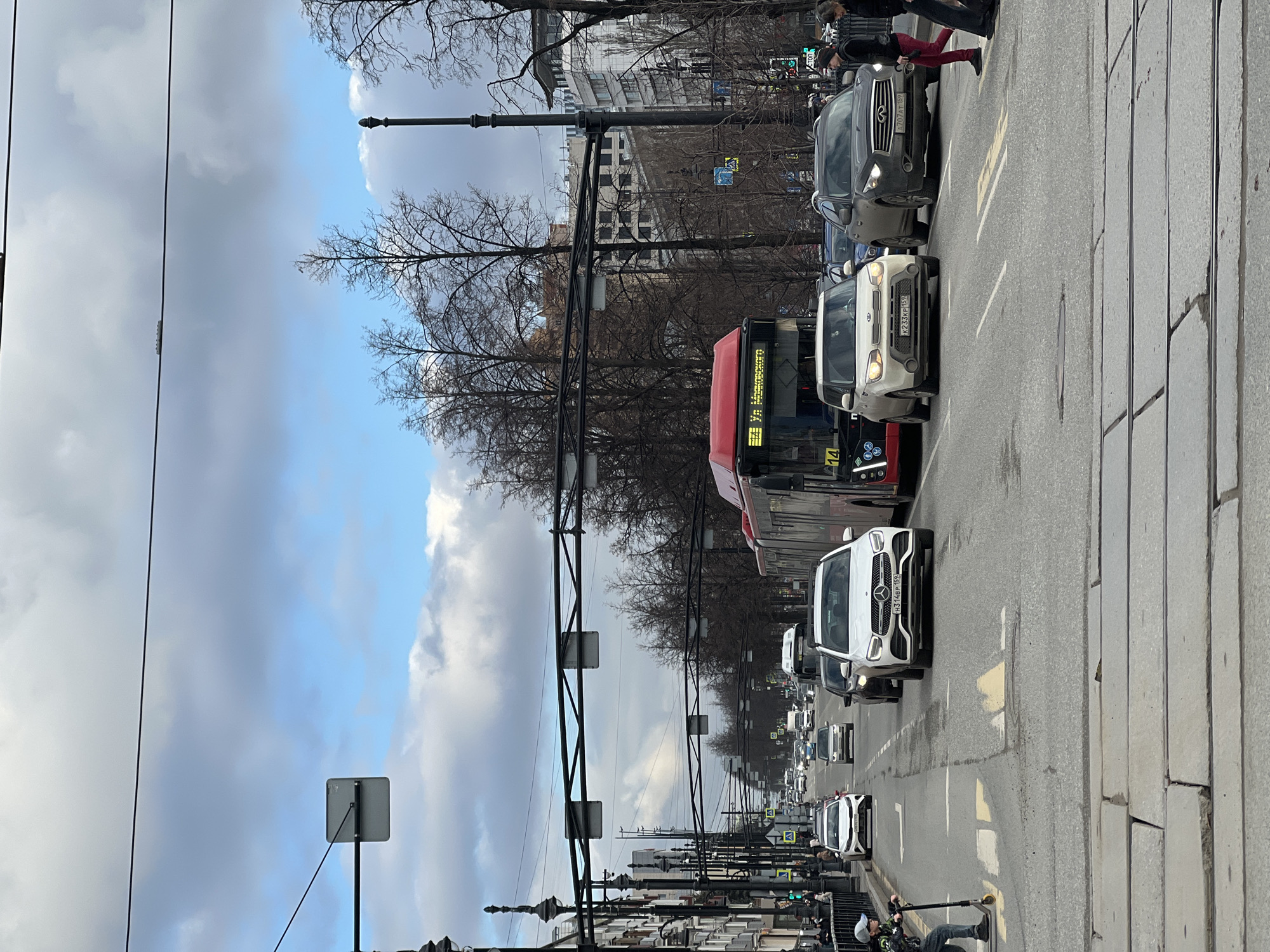 The image depicts a busy street scene in Perm, Russia. A line of cars and a bus are stopped in traffic on a wide avenue paved with stone. Pedestrians can be seen crossing the street near the crosswalk, and others are walking on the sidewalk along the road. Above the street are power lines and a variety of streetlights. Buildings are visible along the road, but they are partially obscured by trees with bare branches. The sky is overcast with a mix of blue and gray clouds.