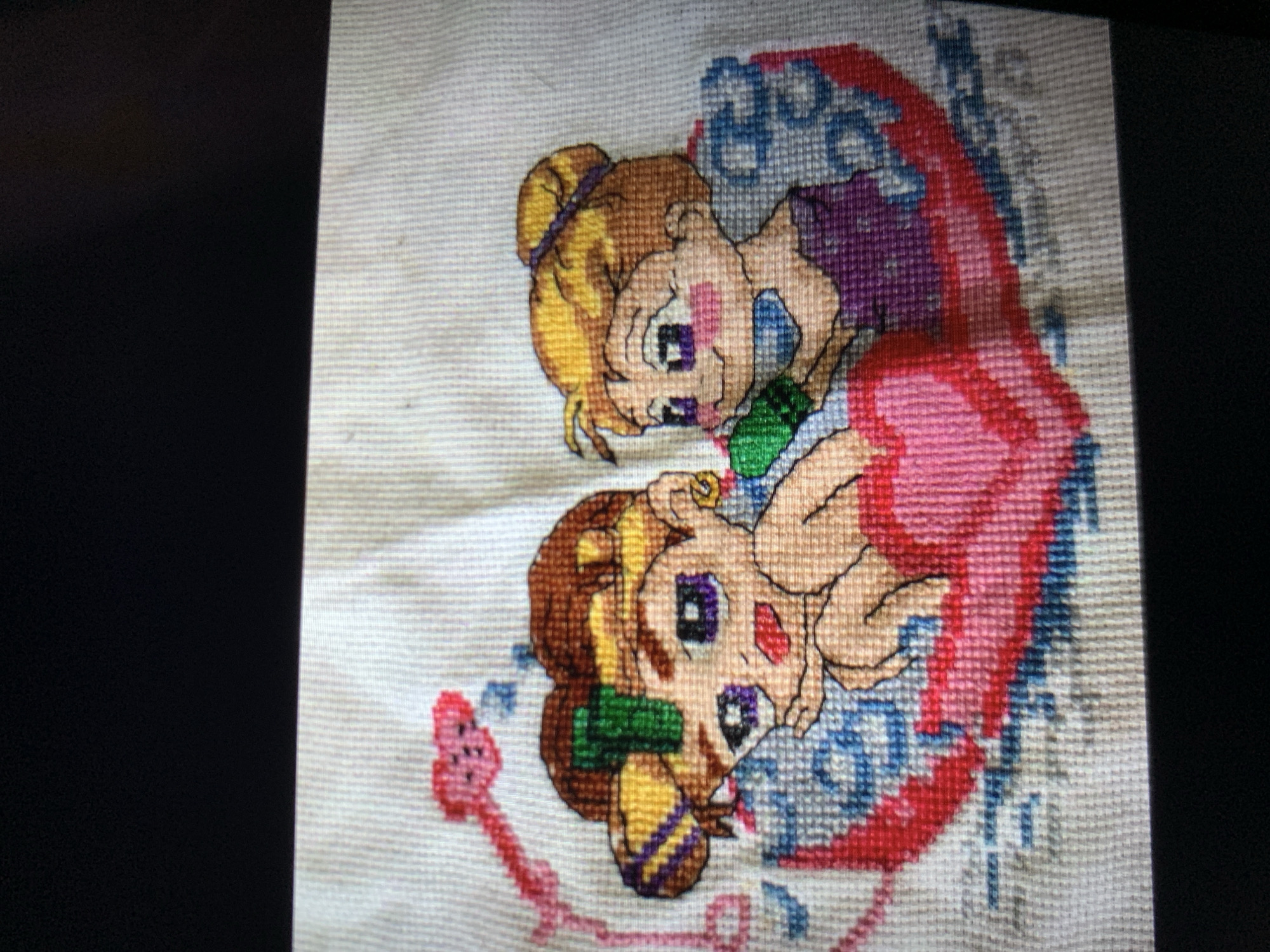 A cross-stitch image depicts two cartoon characters in a heart-shaped object. The character on the left has brown hair, purple eyes, and a green accessory on its head. The character on the right has blonde hair, purple eyes, and is wearing a purple outfit. They are positioned within a large red and pink heart, with blue and white decorative elements around them. A pink heart-shaped object with a stem extends from the upper left of the frame. The background is a white fabric with visible cross-stitch grid lines. The image is a still photograph of the cross-stitch artwork. There is no information provided in the image or crops to determine its location in the city of Lymanka, Ukraine.