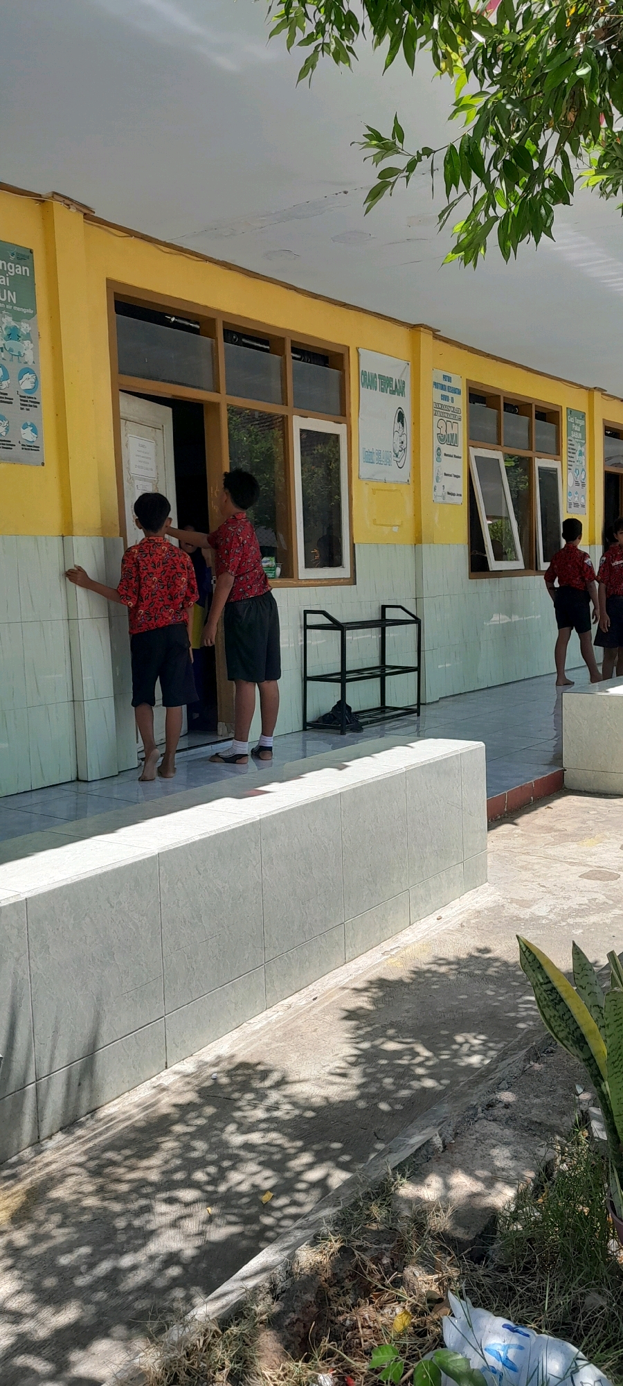 Two young boys, appearing to be students wearing matching red shirts with a pattern and dark shorts, stand on a tiled walkway in front of a yellow building. The boy on the left has his back to the camera and his left hand is on the wall, while the boy on the right is facing slightly towards the building, with his right hand reaching towards the open door. A third student, partially visible in the doorway, appears to be a girl wearing a blue uniform. In the background, a black metal shoe rack is visible. The building has several windows, some open and some closed, and posters are affixed to the yellow wall. The walkway is bordered by a low, tiled wall, and beyond that, the ground is covered with concrete, dry grass, and some scattered debris, including a plastic bottle. The scene suggests an outdoor area of a school, possibly in the city of Rogojampi, Indonesia, given the context. The sunlight casts shadows from overhead foliage onto the ground.