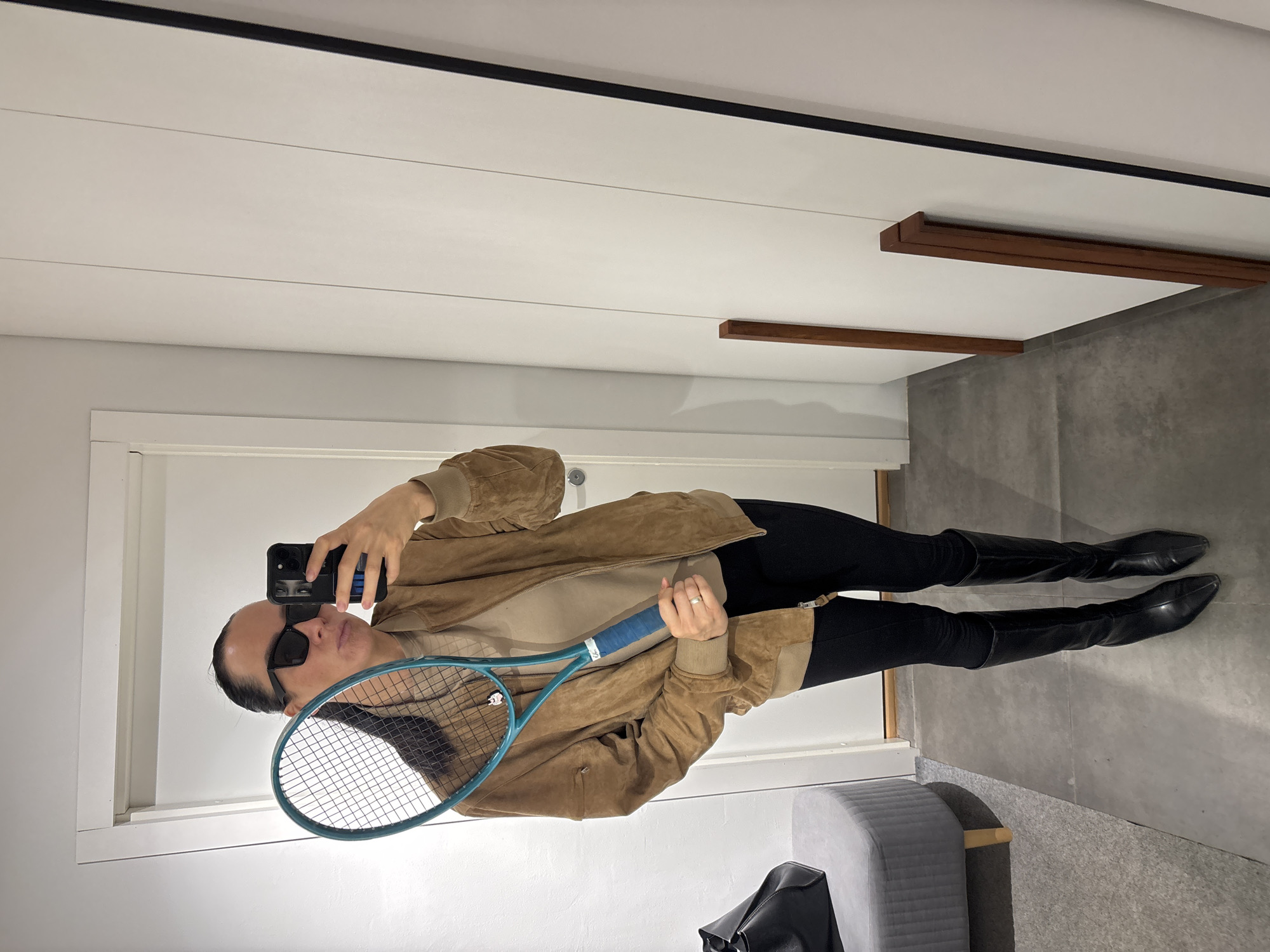 A person wearing black leggings and black knee-high leather boots is standing in what appears to be an indoor hallway or dressing room. They are also wearing a beige sweater and a brown suede jacket. The person is holding a light blue tennis racket in their left hand and a smartphone in their right, apparently taking a selfie in a mirror. The person is also wearing sunglasses. A gray ottoman is visible on the bottom right corner. The ceiling has recessed lighting with black and brown trim. The walls are painted in a neutral color. The location is not discernible from the image.