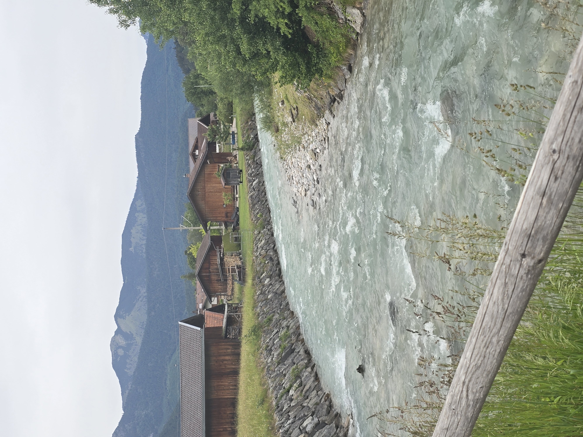 This outdoor image presents a tranquil mountainous landscape featuring a river and traditional buildings, captured under an overcast sky during daytime.
In the midground, a dynamic, light blue-green river flows from right to left, displaying areas of white rapids and visible turbulence. The riverbanks are rocky, giving way to grassy patches and dense green foliage. Along the left bank of the river, several traditional-style wooden buildings, possibly homes or cabins, are nestled into the terrain. These structures feature dark brown or reddish-brown roofs, and some show details such as windows, chimneys, and stacked firewood.
Beyond the buildings, imposing dark blue-grey mountains dominate the background, their slopes covered in extensive forests. The peaks are partially obscured by the overcast conditions. Lush green trees line the river and climb the lower parts of the mountains, contributing to the verdant scenery. On the far right of the foreground, a weathered wooden post, likely part of a fence or railing, is partially visible, surrounded by tall green and dried grasses.
The scene conveys a sense of natural beauty and serenity. There are no people or specific activities depicted. The architecture and mountainous environment are characteristic of the Bavarian Alps, consistent with the Garmisch-Partenkirchen region of Germany. No text is visible in the image.