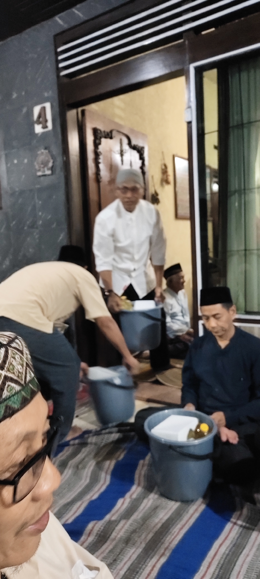 The image depicts an indoor gathering in what appears to be a residential setting in Malang, Indonesia. The scene is dimly lit and the focus is on several individuals, seemingly men, in traditional attire. In the foreground, a man wearing a patterned skullcap and glasses is partially visible, looking to his left. To his right, another man in a dark blue shirt and skullcap is seated, looking down.
Behind them, a man in a white shirt and cap is standing, holding a white object over a blue bucket. Another man in a beige shirt and dark pants is reaching towards a similar blue bucket. In the background, another man in a light-colored shirt and skullcap is seated. The environment includes a wooden door frame, a wall with a decorative element and the number "4" displayed, and striped floor coverings. The overall atmosphere suggests a communal event or religious gathering.