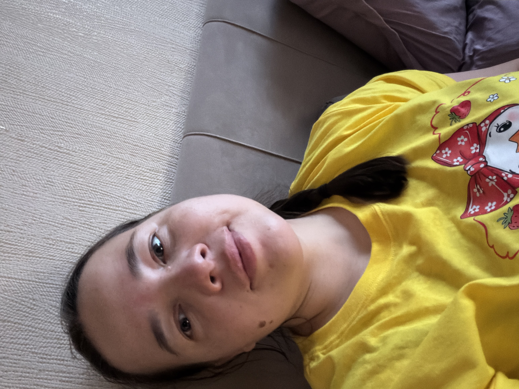 A woman is lying on a couch. The couch has a textured beige section and a grey section. A purple cushion is also visible. The woman is wearing a yellow t-shirt with a cartoon graphic. Her hair is pulled back. It is not possible to determine the location is Chaikovsky, Russia from the image.