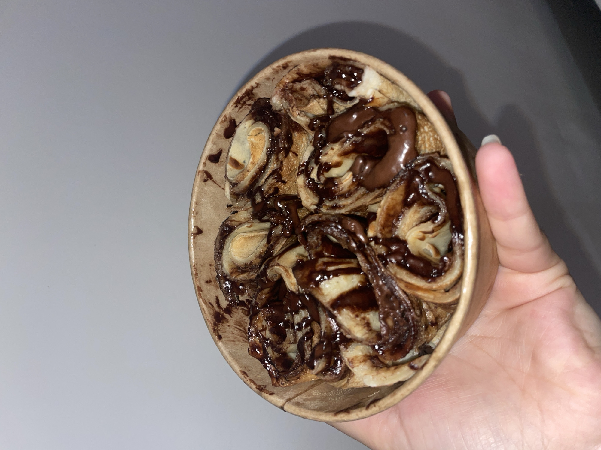 The image shows a paper cup containing small, rolled pancakes or crepes drizzled with a dark chocolate sauce, held in a person's hand. The location is not discernible from the image.