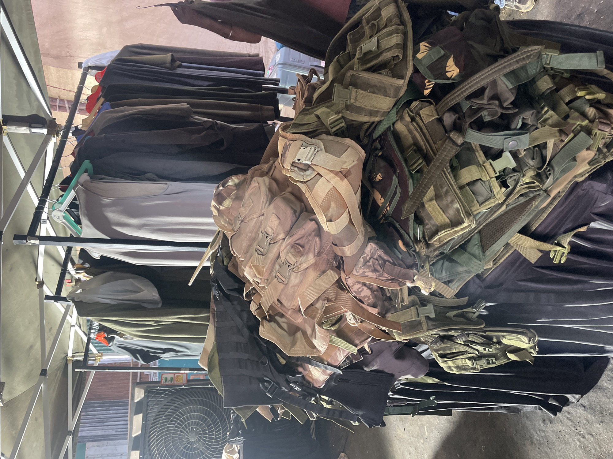 The image shows a stall under a pop-up canopy with clothing hanging on a rack on the left and a pile of military-style tactical vests and pouches on the right. The clothing rack is filled with garments in various shades of gray, brown, and black. The pile of tactical gear is in various colors including tan, olive green, and camouflage patterns. An electric fan is visible beneath the stall. The location is likely in Mira-Bhayander, India.