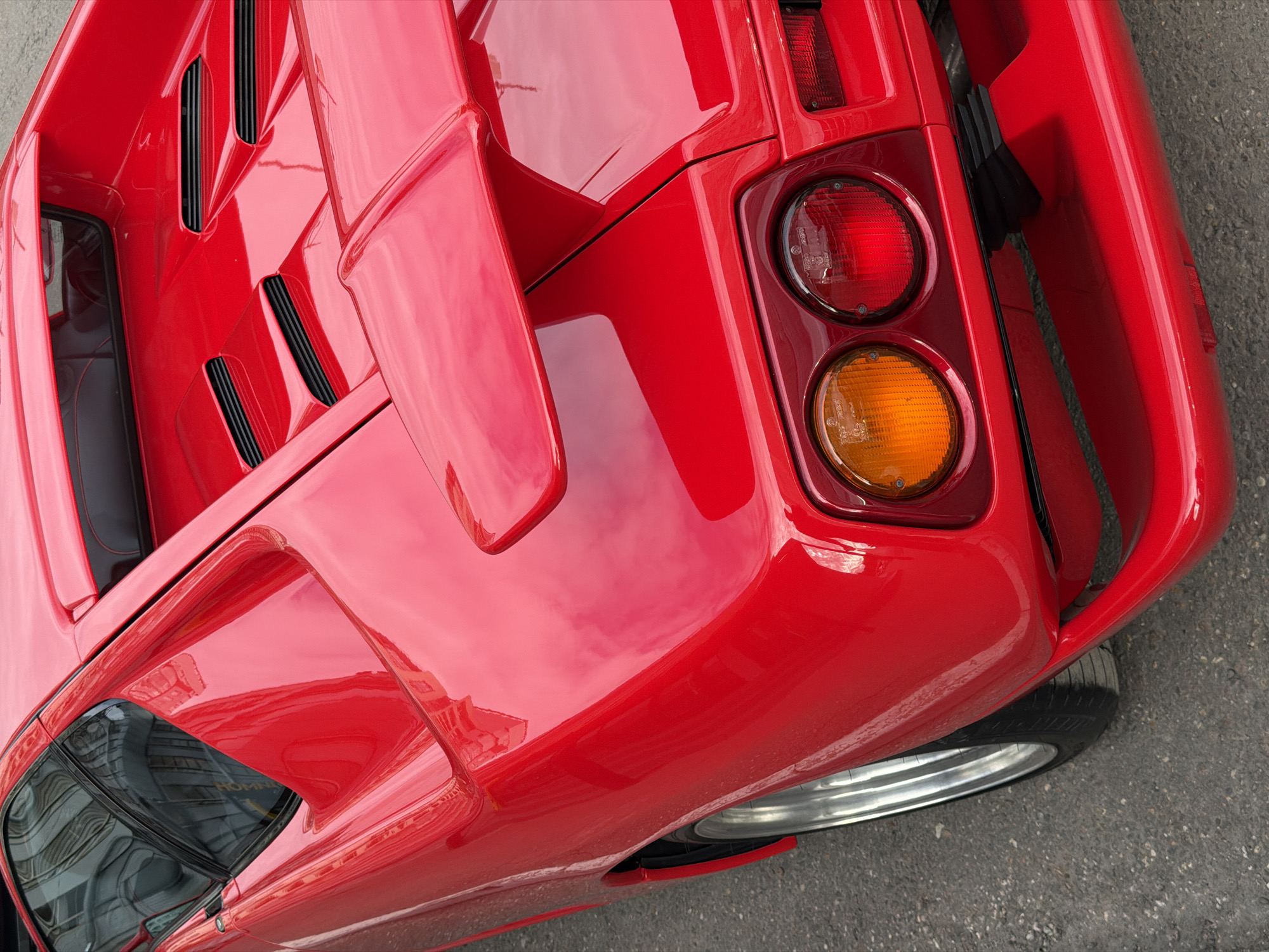 The image presents a close-up, high-angle view of the rear-right quarter of a red Lamborghini Countach sports car, situated outdoors in Moscow, Russia. The vehicle's vibrant red paint is highly polished and reflective, mirroring its surroundings, which appear to be urban structures or sky.
Key visible subjects include the car's distinctive angular bodywork, featuring sharp lines and geometric forms characteristic of the Countach model. The rear deck incorporates black louvers for engine cooling and a prominent, angular spoiler-like element. The car's taillight assembly consists of two vertically stacked circular lights: an upper red light and a lower amber light.
A portion of a silver multi-spoke wheel and a black tire are visible at the bottom right, resting on asphalt pavement. The car appears stationary, suggesting it is parked or on display. Reflections on the side window/vent area reveal distorted, partially legible text, which could be "HORN" or similar, hinting at a nearby sign or building. The scene takes place during the daytime, under clear weather conditions, enhancing the car's striking color and pristine condition.