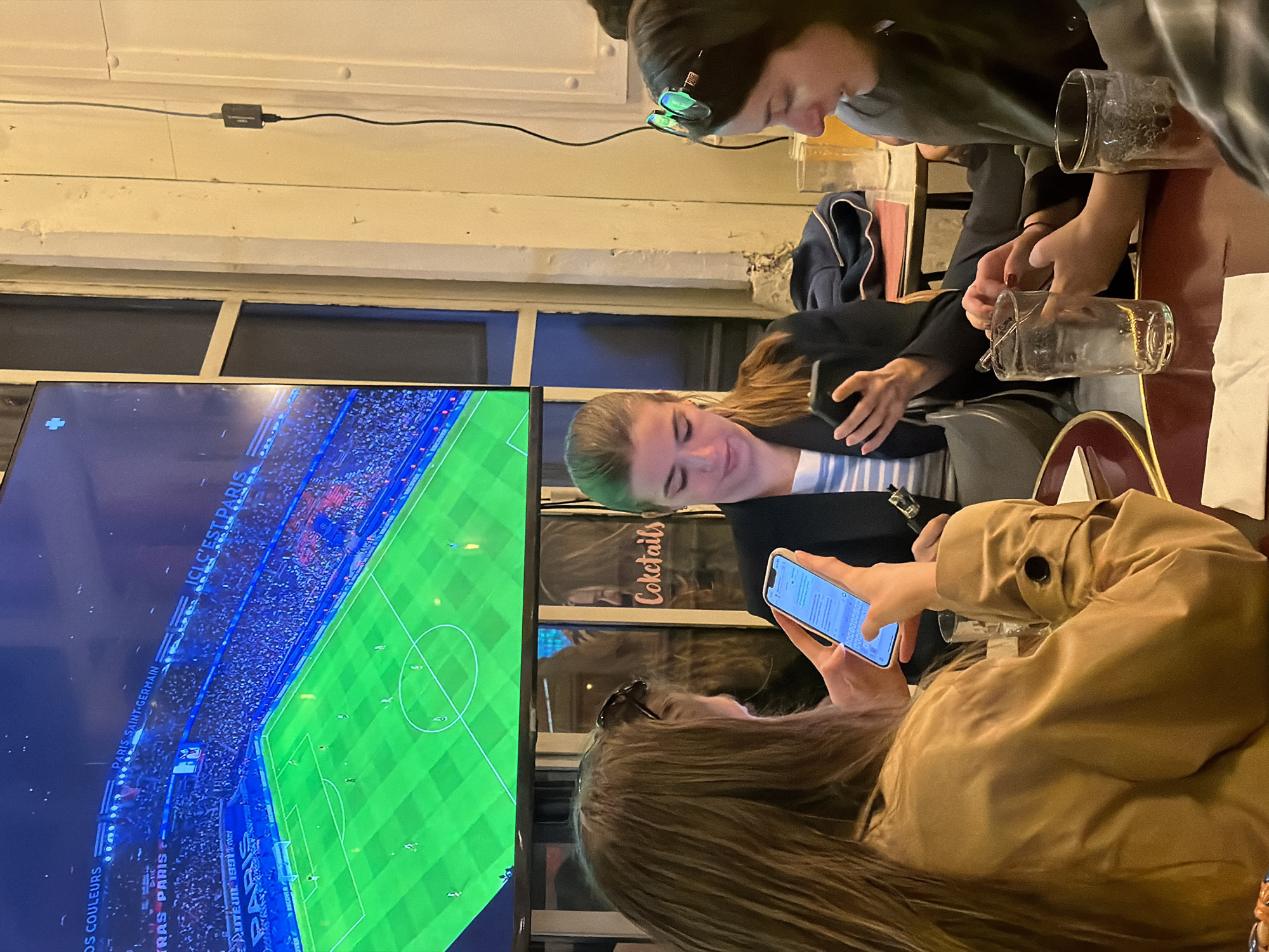 Three people are seated at a table indoors. A woman with long brown hair is holding a smartphone and looking at its screen. A woman in a blazer is looking at a smartphone that is being held by a person out of frame. A person with sunglasses on their head is sitting next to her. Empty glass beer mugs are on the table. A large television screen is showing a soccer game, identifying the team Paris Saint-Germain, which is based in Paris, France.