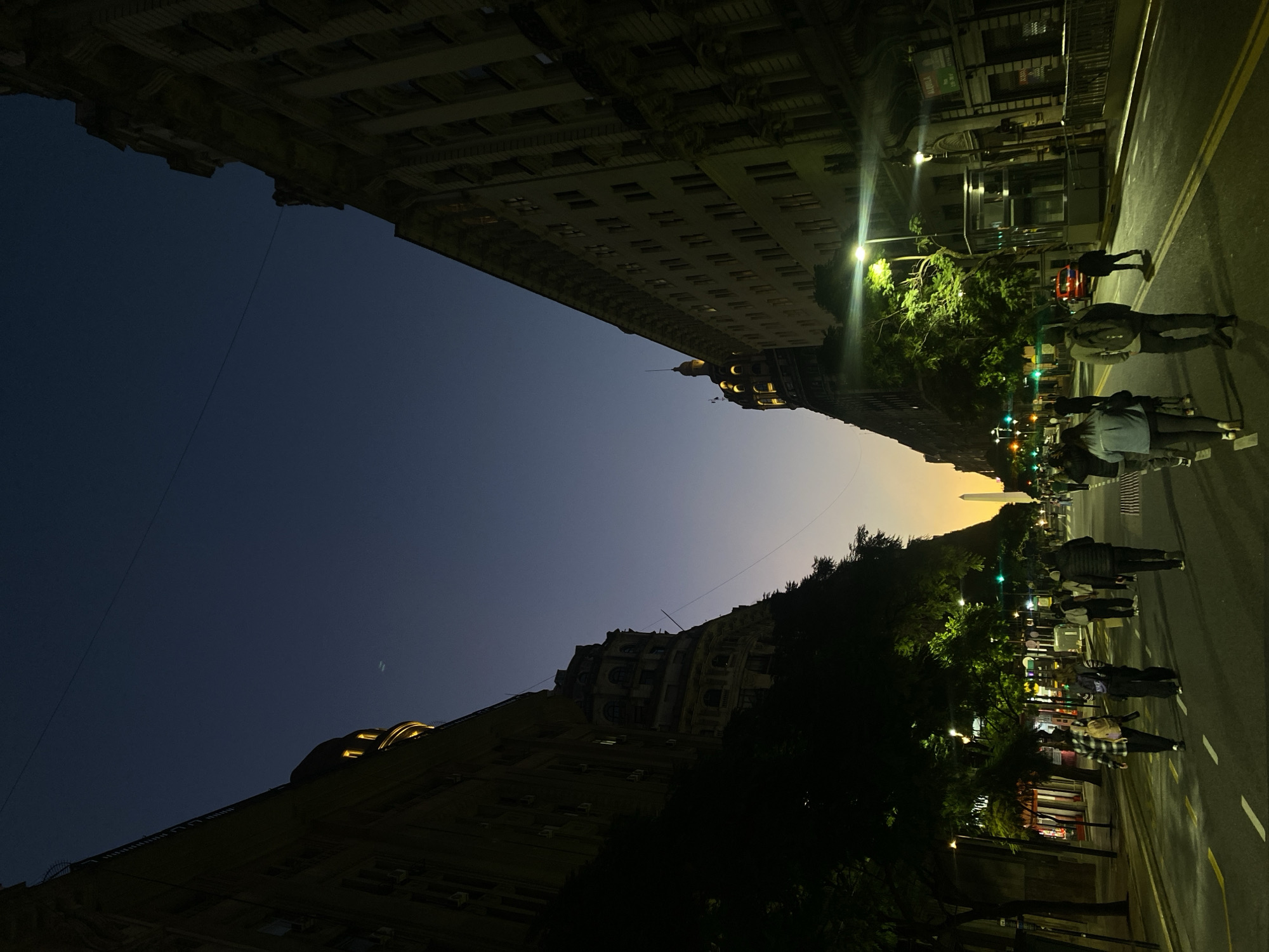 The image captures a nighttime street view in Buenos Aires, Argentina, looking upwards between tall buildings. The sky transitions from dark blue to a faint orange glow near the horizon. On the right side of the image, streetlights illuminate a pedestrian crosswalk and a line of people walking along the sidewalk. Trees are visible alongside the street. In the distance, the obelisk of Buenos Aires stands as a prominent landmark. The buildings on both sides are dark but show windows and architectural details. The scene suggests an urban environment with active pedestrian traffic in the evening.