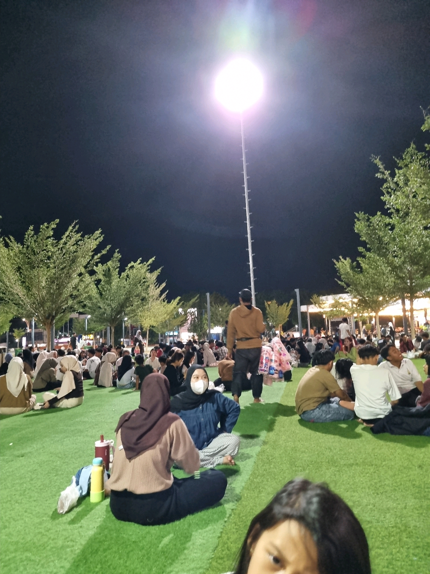 A nighttime scene depicts a gathering of people seated on a bright green, artificial turf lawn in what appears to be a public park or event space. A tall, bright light pole with a powerful lamp illuminates the area, casting a glow upwards and creating lens flares. The sky is dark, indicating it is nighttime. Numerous individuals, including men, women, and children, are dispersed across the lawn, some sitting individually and others in small groups. Many of the women are wearing hijabs. A few people are standing. In the background, there are trees with green foliage, and further back, structures that could be shops or food stalls are visible, adorned with string lights. In the foreground, a person's head and shoulders are visible, partially obscuring the view of the scene. Two water bottles and some clothing items are placed on the grass near the foreground subjects. The overall impression is one of a casual outdoor event, possibly a market or festival, taking place in Tanamodindi, Indonesia.