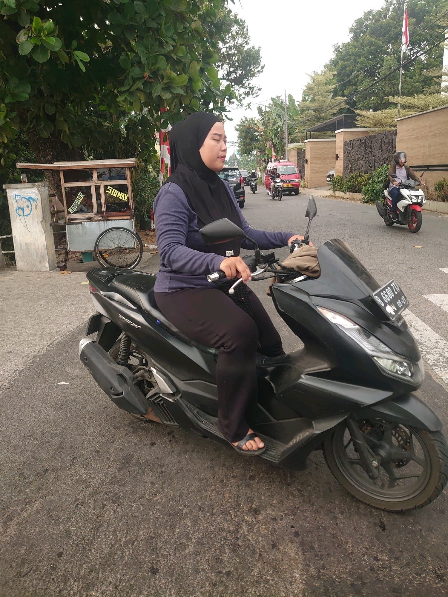 In Depok, Indonesia, a woman wearing a black hijab and a blue shirt is riding a black scooter. She is wearing black pants and sandals. The scooter has a license plate that reads "B 6880 ZTQ". The woman is looking to her right. In the background, there is a street with other vehicles and buildings. To the left of the woman, there is a food cart with a bicycle wheel. A Indonesian flag is visible on a pole behind a wall. The sky is overcast.
