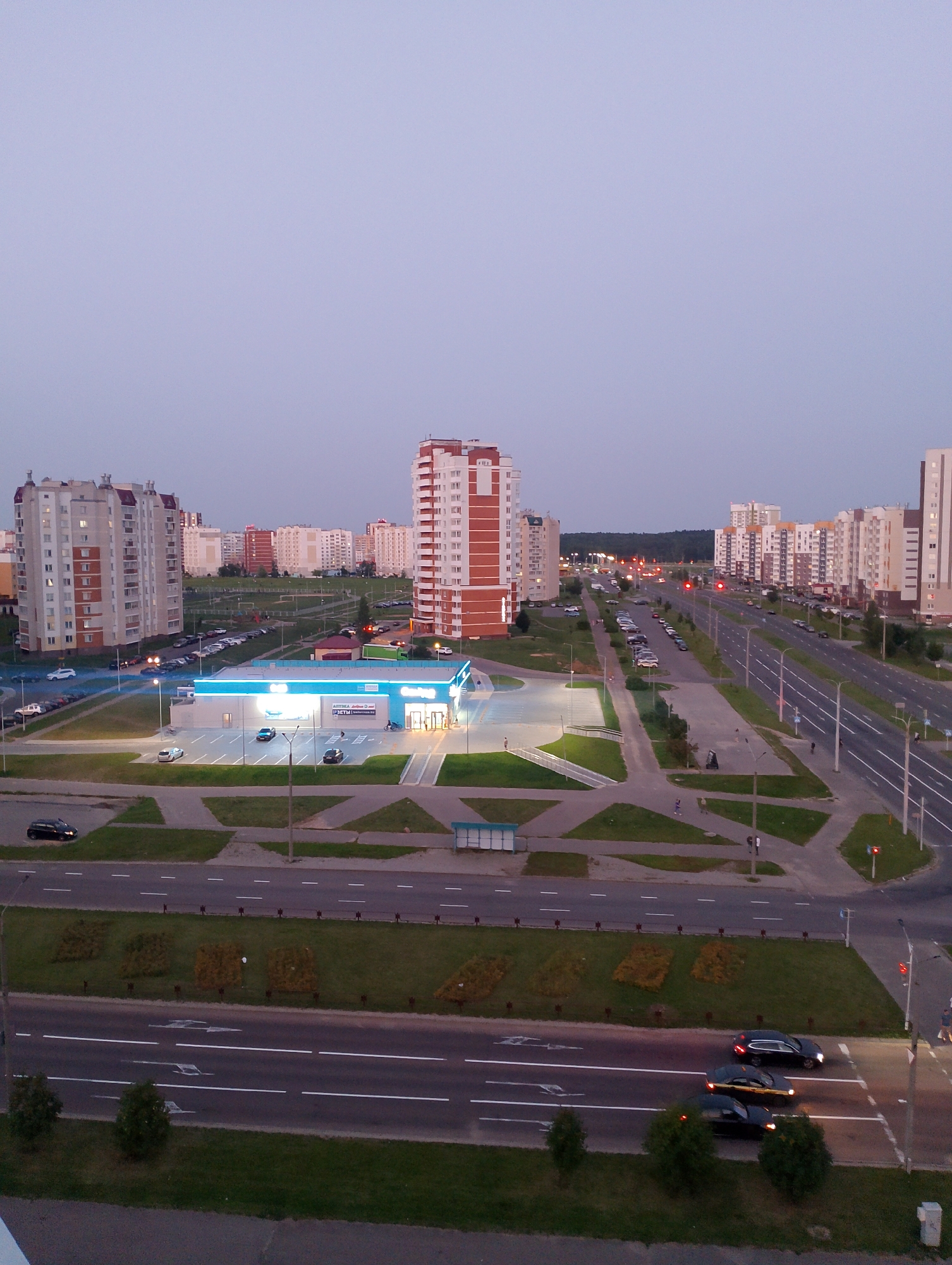 The image captures an aerial view of a city intersection at dusk, likely in Vitsebsk, Belarus. In the foreground, a multi-lane road runs horizontally, with several vehicles in motion. To the left of this road, a grassy median is visible with patterned landscaping and a lamppost. Further back, a brightly lit supermarket with a blue awning and illuminated signage stands prominently in the center of the frame. Surrounding the supermarket is a parking lot with parked cars. Beyond the supermarket, a series of multi-story residential buildings, characterized by their light-colored facades and red accents, are visible. Another, taller, red-brick building with numerous windows is situated to the right of the supermarket. In the background, more buildings and a line of trees suggest the edge of the urban area. The sky is a uniform, muted blue, indicating twilight. The scene is well-lit by the artificial lights of the buildings and streetlights.
