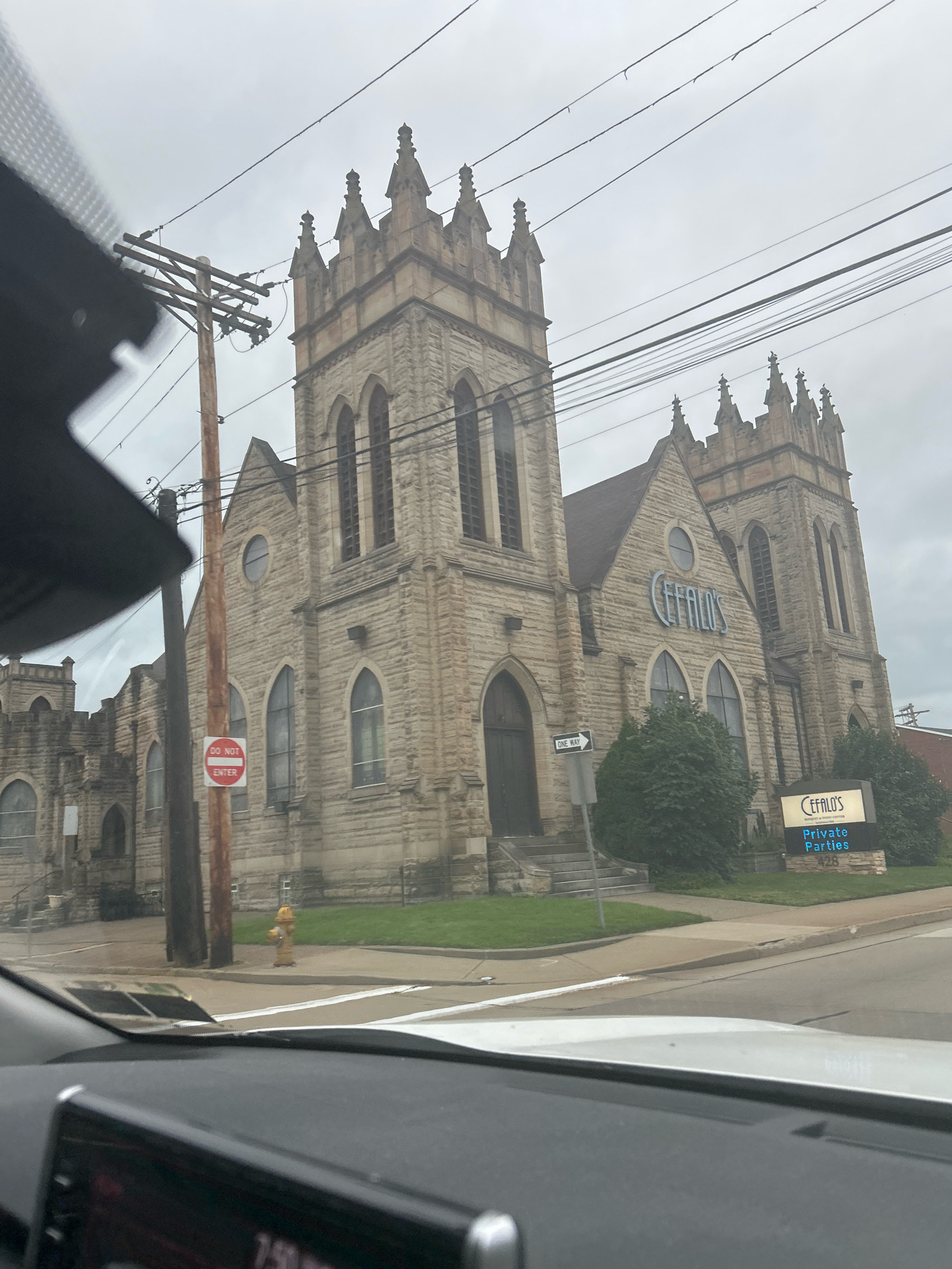 An exterior medium shot captured from a vehicle shows the Cefalo's wedding and reception venue, formerly a church, in Carnegie, Pennsylvania, on an overcast day. The historic stone building features Gothic architectural elements such as pointed arch windows and crenellated towers. A prominent blue neon sign on the facade reads "CEFALO'S." Below this, a smaller lit sign indicates "Private Parties" with the address "428." A wooden utility pole with wires stands to the left of the building, and a red and white "DO NOT ENTER" sign is affixed to it. A yellow fire hydrant is visible on the sidewalk in front of the building. The foreground is dominated by the interior of a car, including a dashboard and windshield.
