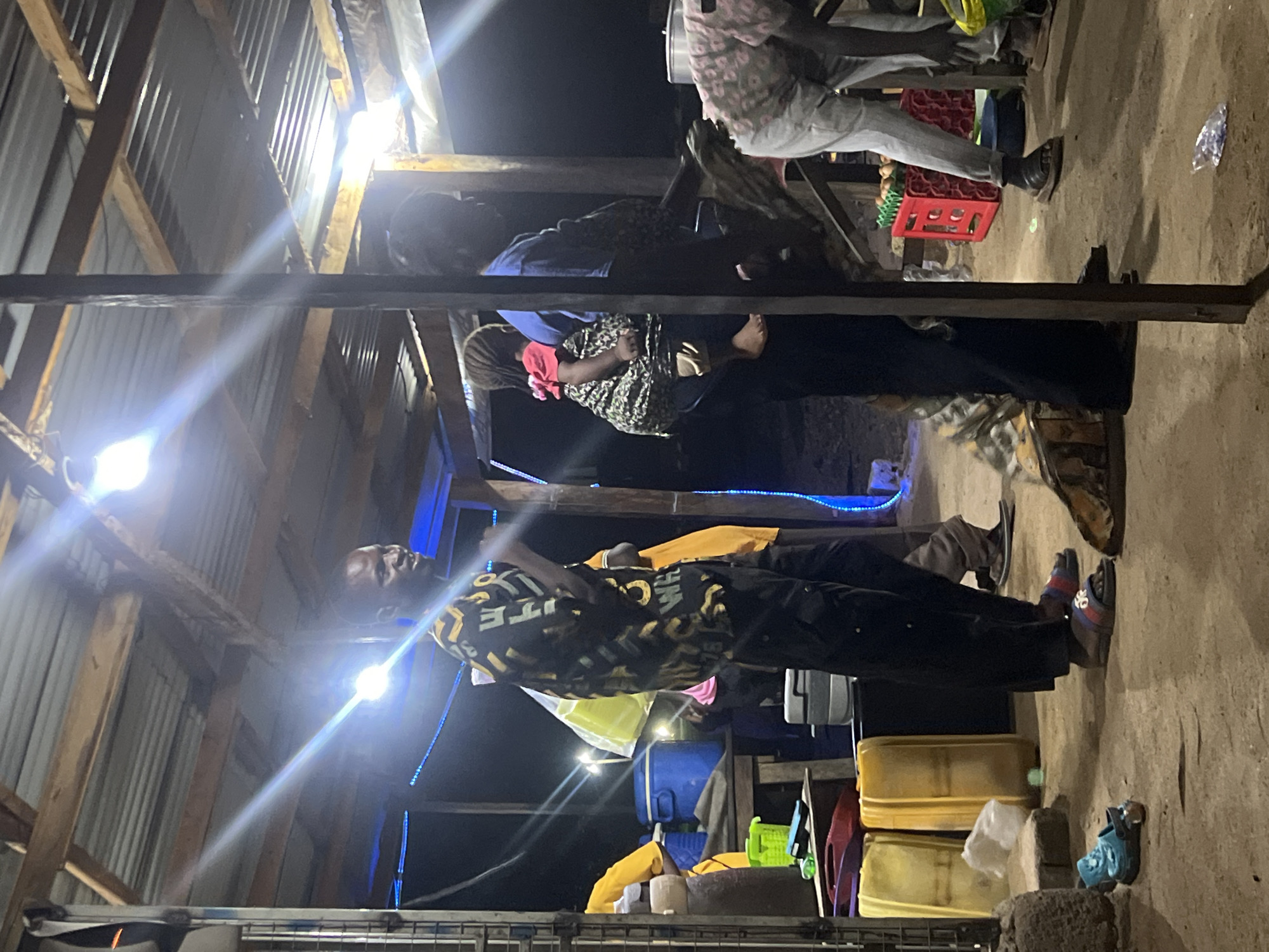 The scene appears to be taken in a makeshift outdoor space in Jalingo, Nigeria. The location is lit by bright overhead lights attached to the wooden frame of a structure with a corrugated metal roof. Several people are present, though some are partially obscured or out of focus. One man stands prominently in the foreground, wearing a patterned shirt and dark pants. Other individuals are visible, including a person in a red shirt and several others gathered under the wooden beams.
Various objects are scattered around the ground, including plastic containers, a crate, and personal belongings like shoes. The ground appears to be dirt or packed earth. Overall, the setting suggests a casual gathering in a semi-outdoor, functional space, likely a roadside or market area.