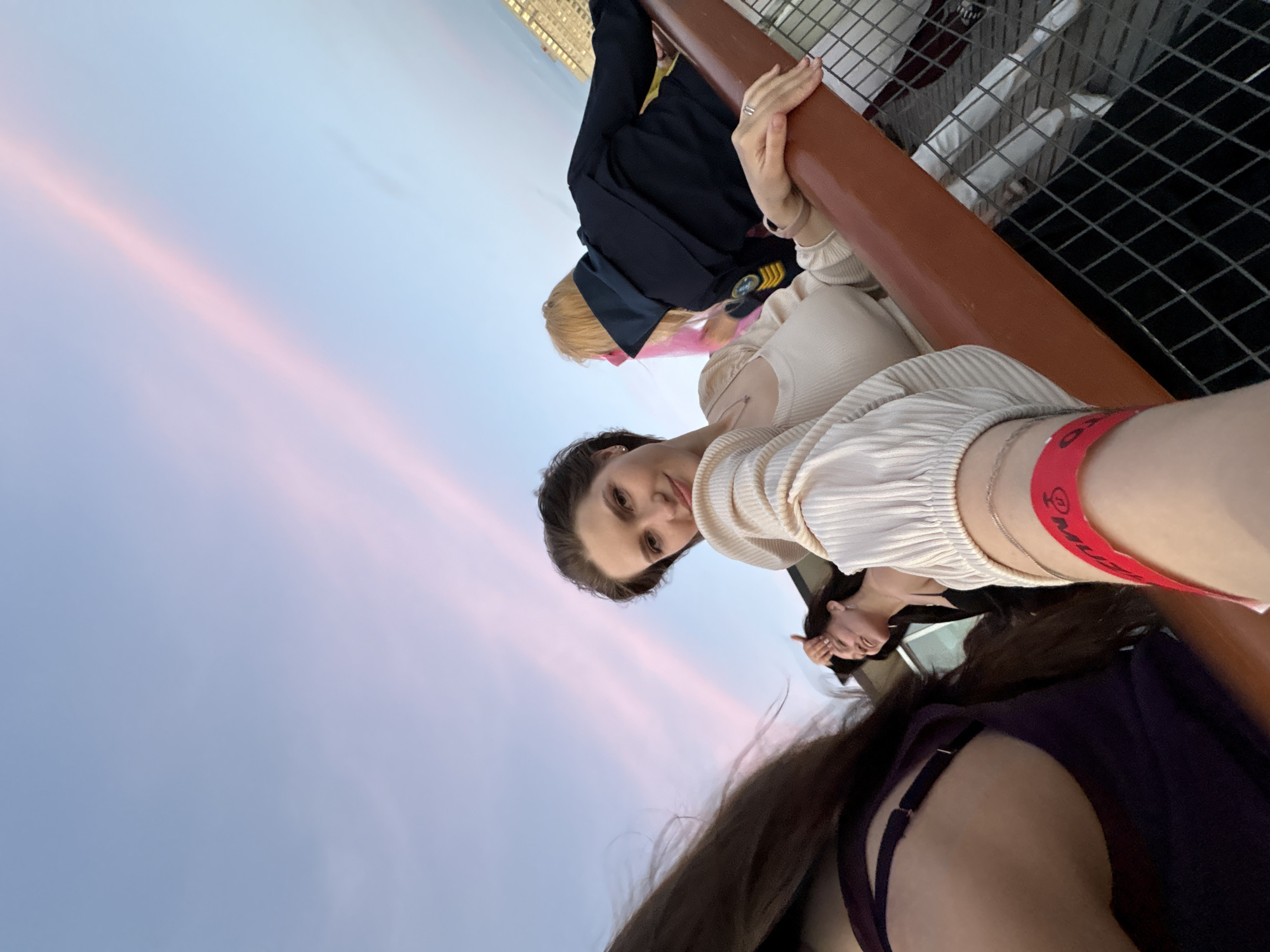 A group of women are gathered on an outdoor deck or balcony with a view of the city of Saint Petersburg at dusk. The sky is a gradient of soft blues and pinks, with thin streaks of pink clouds. In the foreground, a woman with dark hair pulled back smiles at the camera, wearing a cream-colored, ribbed top with puffed sleeves. A red wristband is visible on her arm. To her left, another woman with long dark hair, wearing a dark purple top, is partially visible. To the right of the main subject, a woman with blonde hair, wearing a dark jacket, leans on a railing. In the background, buildings with lights are visible against the darkening sky. The railing is made of dark wood, and a wire mesh fence is visible below. The scene appears to be from a rooftop bar or observation deck, possibly in the historic center of Saint Petersburg, given the architectural style of the background buildings. The atmosphere suggests a social gathering or event.