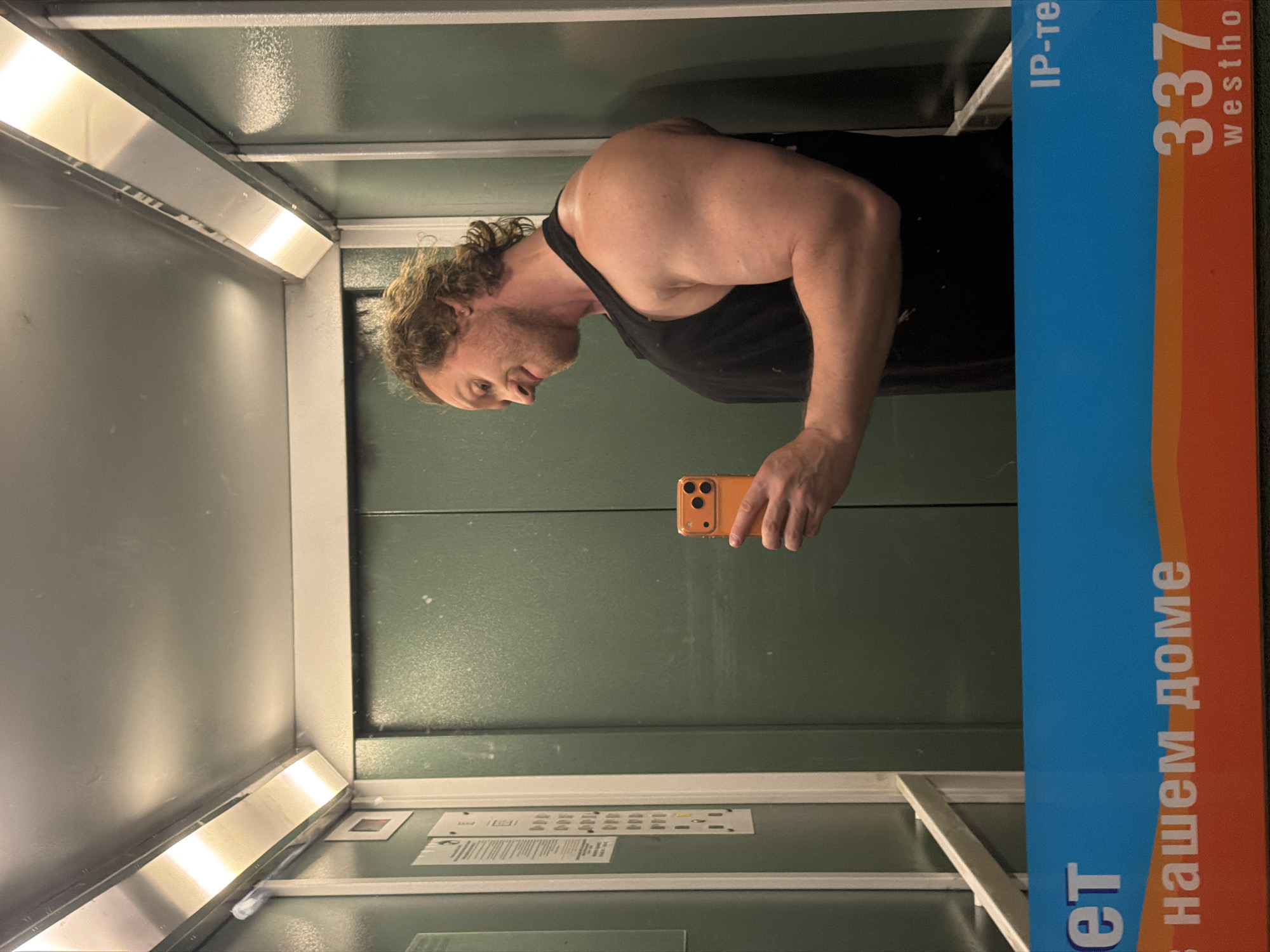 An adult male with light brown curly hair and a beard, wearing a black sleeveless tank top, is captured in a selfie reflected in the mirrored surface of an elevator. He holds an orange smartphone in his right hand, positioned to reflect his image. He exhibits a subtle smirk and a muscular physique, particularly his right arm and shoulder, which are prominently displayed. The elevator interior features light green walls with visible smudges, a light grey ceiling with two illuminated rectangular light fixtures, and a control panel on the left wall with multiple buttons and a red indicator light. A blue and orange banner is present at the bottom of the reflected scene, displaying Cyrillic text including "нашем доме" (in our house) and "IP-те", along with "337" and "westho" (likely an abbreviation for Westhome, a known internet provider in the region). This combination of Cyrillic text and the Westhome branding places the location in Saint Petersburg, Russia.