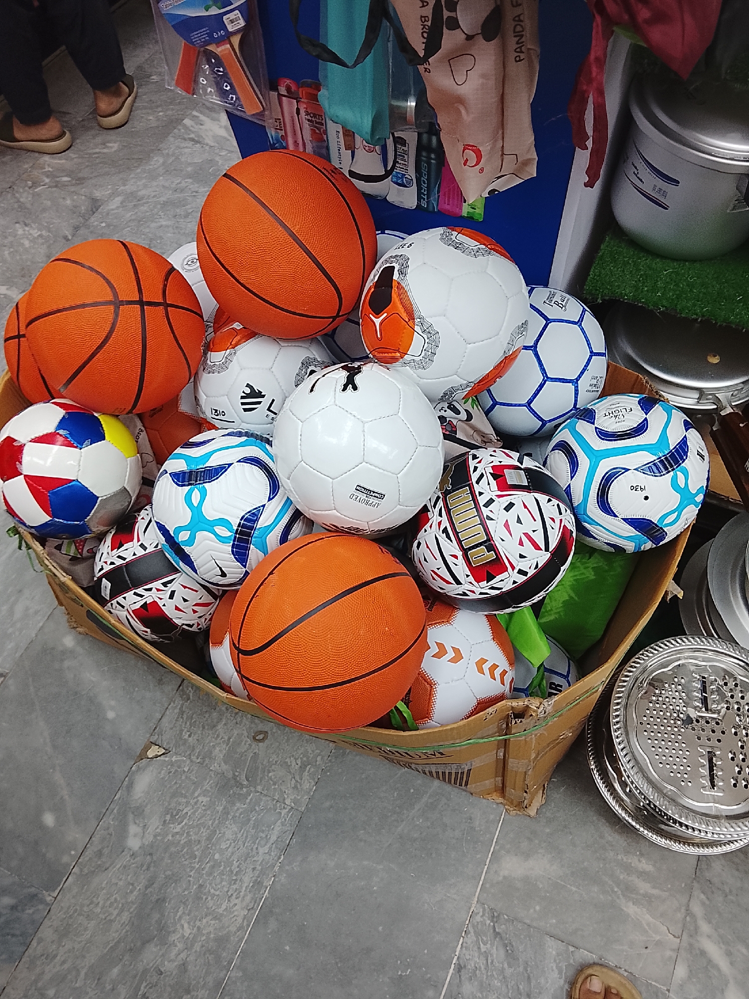 The image shows a cardboard box filled with basketballs and soccer balls. The box is placed on a gray tiled floor in what appears to be a shop, possibly in Gulgasht Colony, Pakistan. There are three orange basketballs and several soccer balls with various designs. To the right of the box, there are some metal kitchenware items. In the background, there are items on a shelf, including kitchenware and sports items. Parts of two people's feet are visible in the background and foreground.