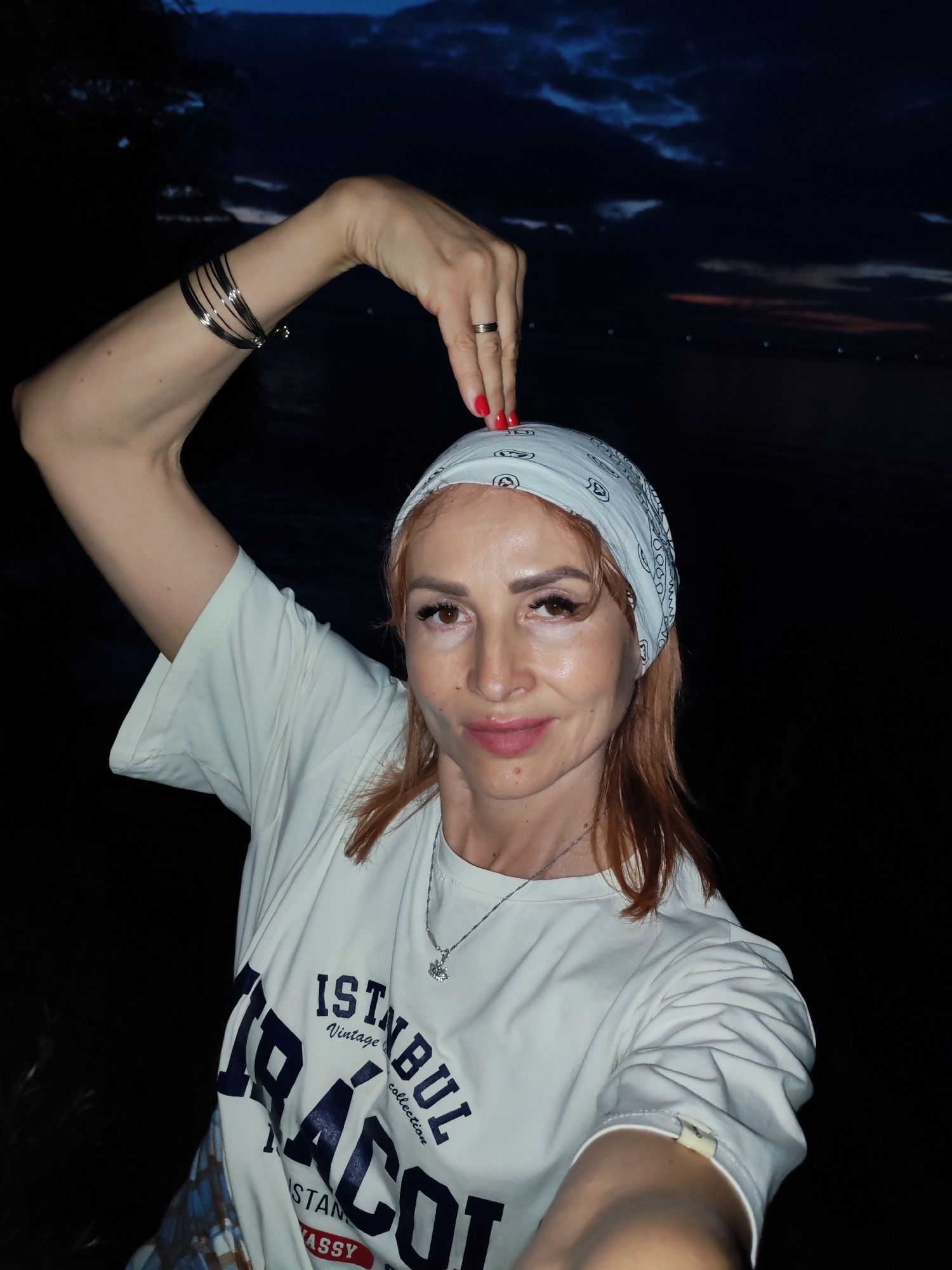 A woman with auburn hair is shown from the chest up, posing for a selfie at dusk near the Kama River in Chaikovsky, Russia. She is wearing a white t-shirt with "ISTANBUL Vintage collection" printed on it, a white bandana tied around her head, and several silver bracelets on her left arm. Her left arm is raised above her head, with her hand in a gesture over the bandana. Her right hand is out of frame, likely holding the camera. The background shows a dark sky with clouds, indicative of twilight, and the calm waters of the Kama River.