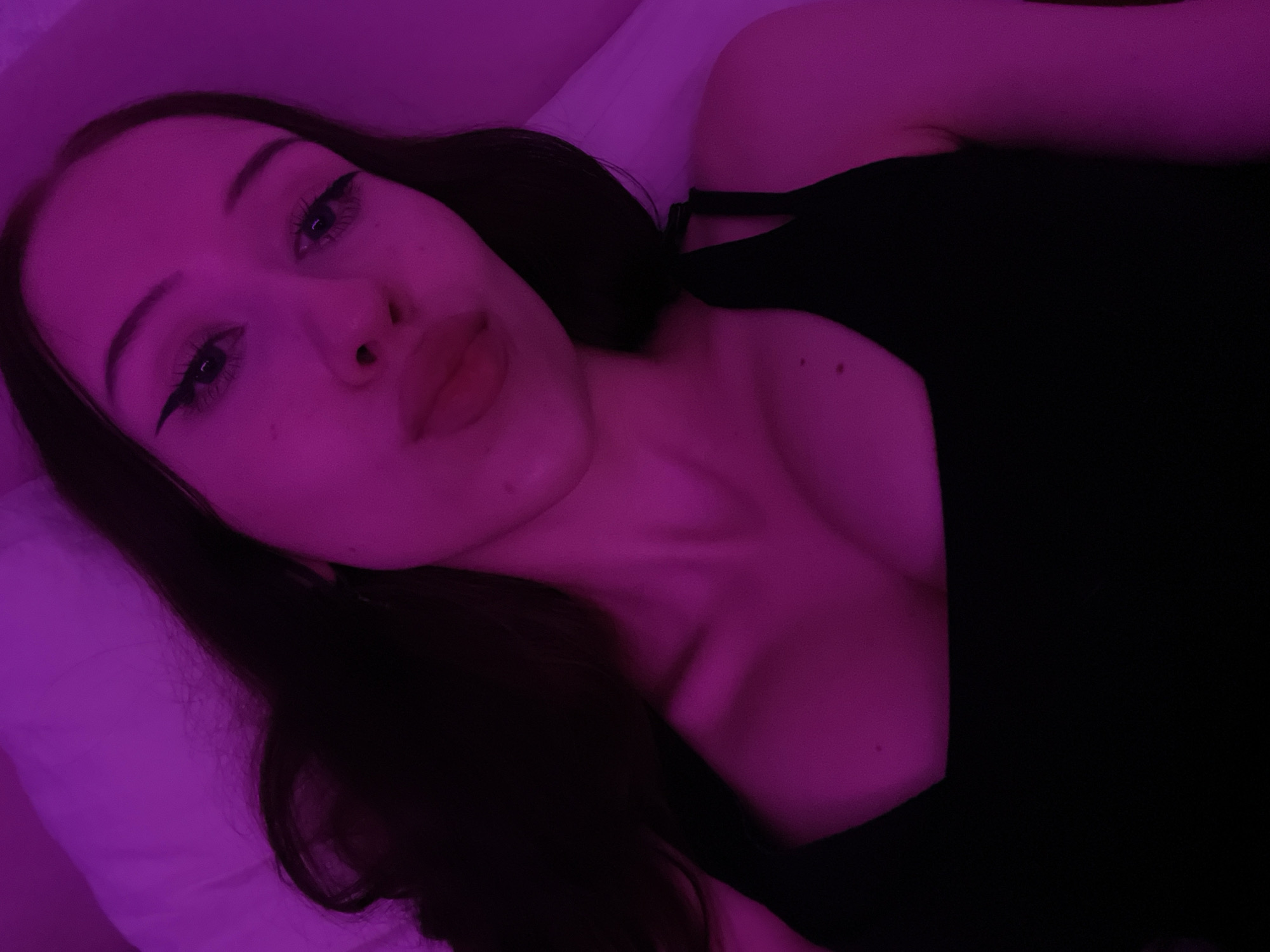 The image shows a close-up, waist-up shot of a young woman with dark hair and full lips, wearing a black tank top. She is illuminated by a pink light. The background consists of a white pillow and appears to be a bedroom setting. There are no discernible landmarks or features that would allow for the specific identification of a location within the city of Votkinsk, Russia.