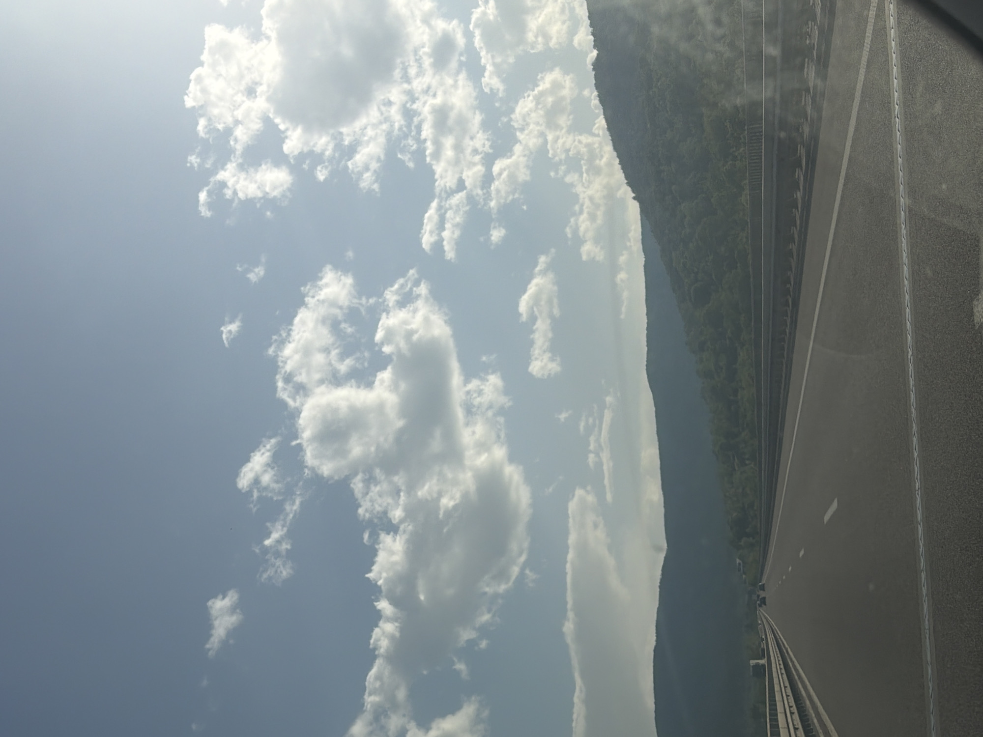 The image shows a highway near Dolno Varshilo, Bulgaria, viewed from the inside of a vehicle. The asphalt road surface has visible lane markings. A mountain covered in dense trees borders the road on one side, while a blue sky with scattered white clouds is visible above. The view suggests motion along the highway.