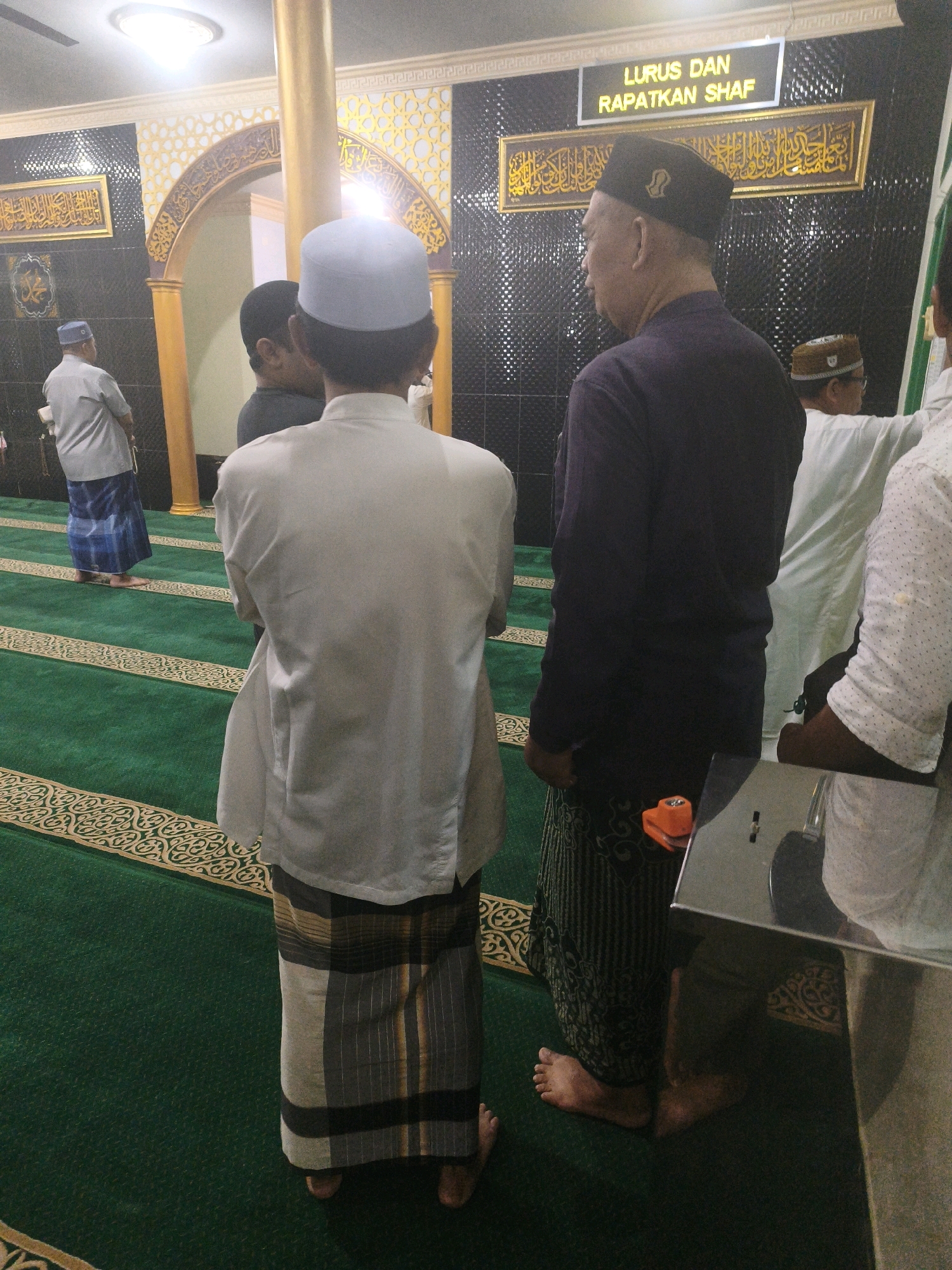 The image depicts an interior scene, likely a mosque, in Samarinda, Indonesia. The floor is covered in a green carpet with decorative gold patterns. Several men are present, all dressed in traditional Islamic attire, including sarongs and skullcaps.
In the foreground, a man wearing a white skullcap and a white garment is seen from behind, facing away from the camera. To his left, another man in a dark skullcap and a dark purple shirt, also viewed from behind, stands with his arms at his sides. He is wearing a patterned sarong. To the far right, partially visible, is a man in a white shirt with a pattern of small dots. In the background, another man in a gray shirt and a blue and white patterned sarong is walking away from the camera.
The environment appears to be a hall with decorative arches and Islamic calligraphy on the walls. A prominent sign above the men reads "LURUS DAN RAPATKAN SHAF," which translates to "Straighten and Tighten the Rows," a common instruction during congregational prayer in Islam. A golden pillar is visible towards the left of the frame. The lighting suggests artificial illumination. There are no explicit indicators of the specific location within Samarinda, Indonesia, beyond the cultural context implied by the attire and religious instruction.