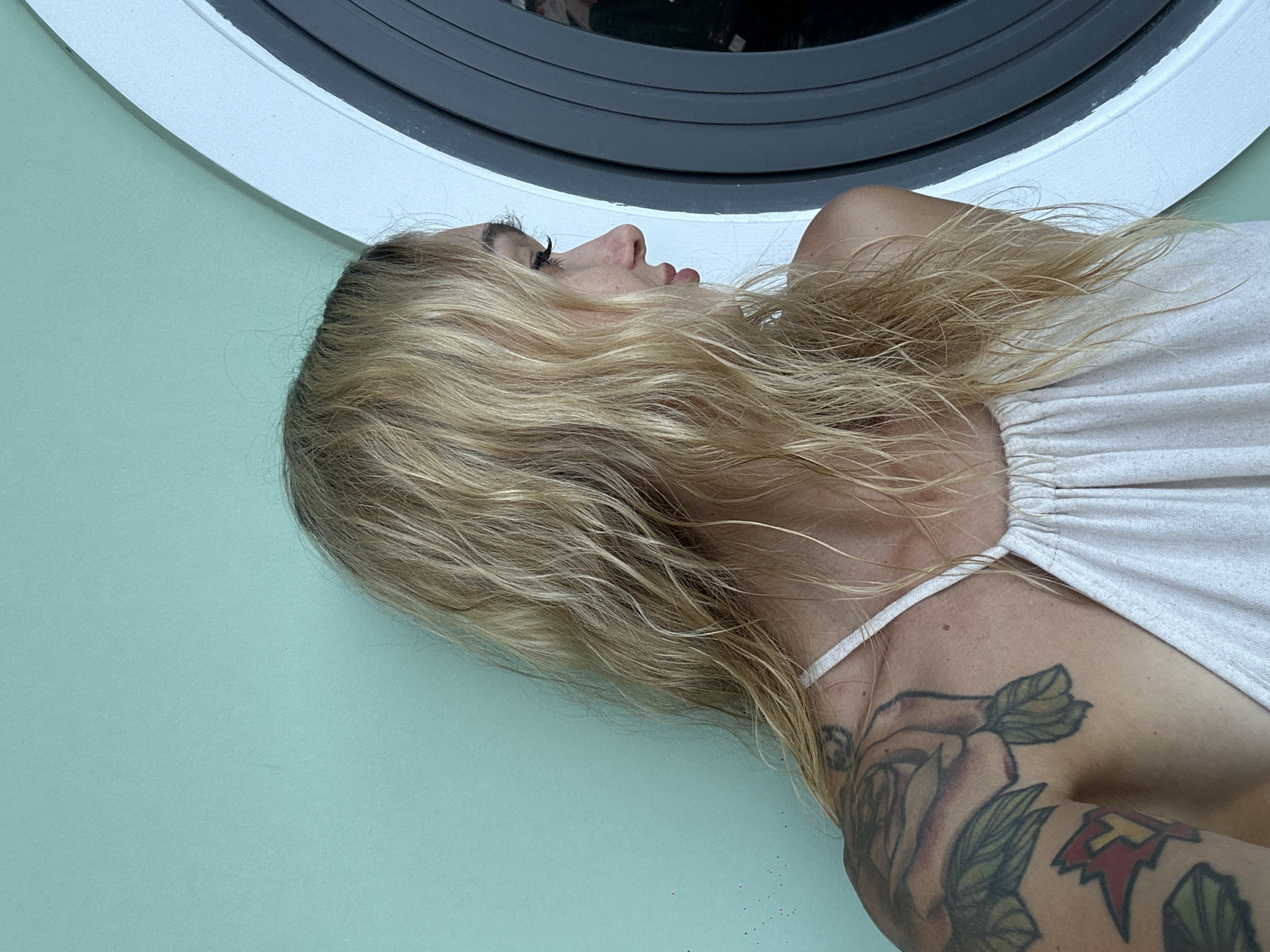 The image presents a close-up, side-profile view of a woman with long, flowing blonde hair, positioned against a plain, light teal or mint green background. She appears to be lying down or reclining, with her head turned to the left and her gaze directed slightly upwards. Her eyes are open, and her expression is neutral and calm. Prominent eyelashes are visible on her closed eye.
She is wearing a light-colored, possibly white or off-white, top with a thin shoulder strap. A large, detailed tattoo of roses and foliage covers her right shoulder and upper arm, with another smaller, geometric-patterned tattoo also visible on her upper arm. Her long, wavy blonde hair is spread out around her head and shoulders, showing variations in tone.
In the upper background, a circular object with a dark inner rim and a lighter outer frame, possibly a window or porthole, is partially visible above her head. The rest of the background consists of a solid, smooth light teal wall. No specific location cues for Mũi Né, Vietnam, are present in the image itself. No text is visible. The scene conveys a sense of quiet repose.