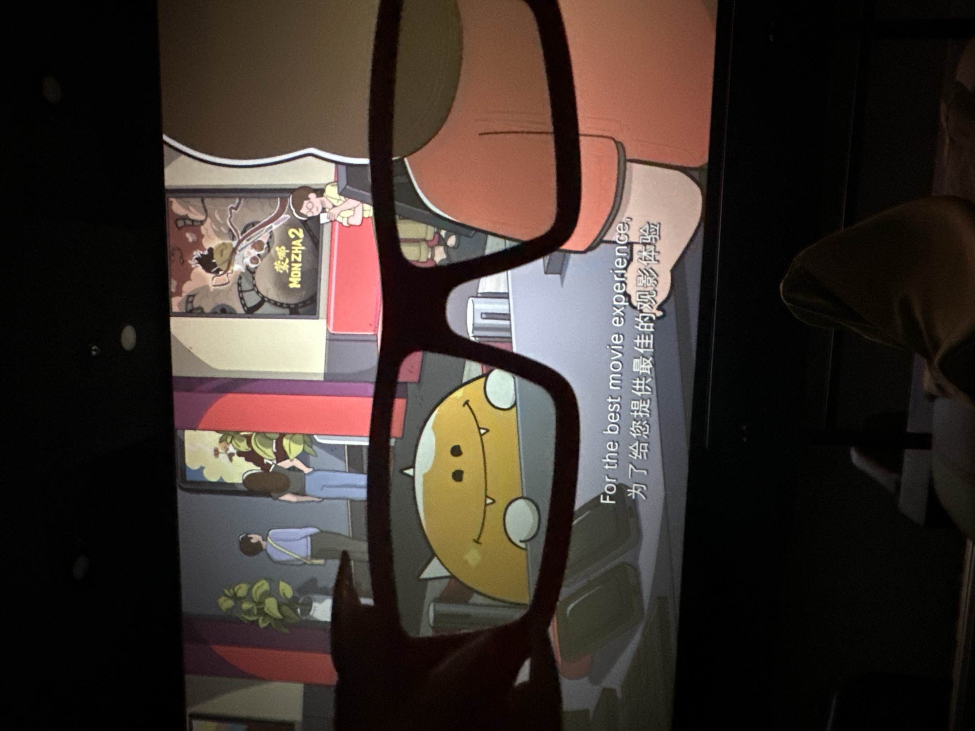 A person is holding a pair of red-rimmed 3D glasses up to a movie screen. The glasses frame a cartoon character that appears to be yellow and round with a large smile and black dots for eyes. In the background of the screen, two figures are standing near plants, and a movie poster is visible. Text on the screen reads "For the best movie experience," and in Chinese characters, "为了给您提供最佳的观影体验." The scene appears to be in a movie theater. The city of Khan Daun Penh, Cambodia is not depicted in this image.